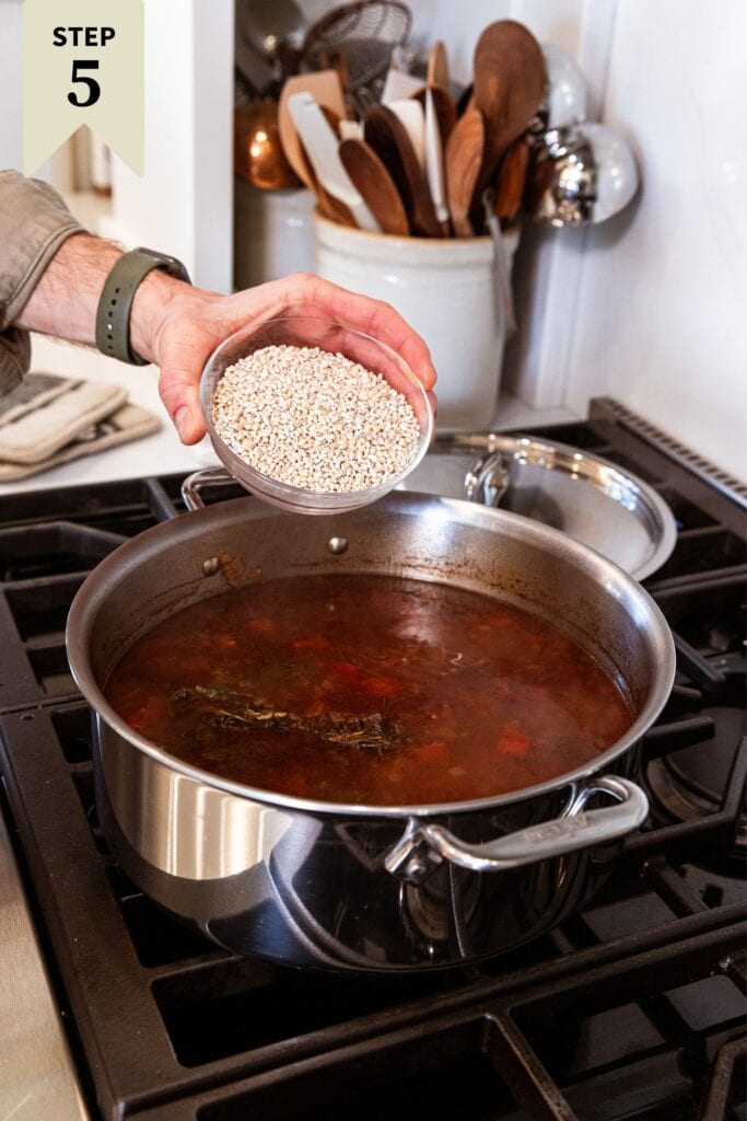 Hand about to pour pearled barley from glass bowl into large metal stockpot filled with cooking brown-colored soup.