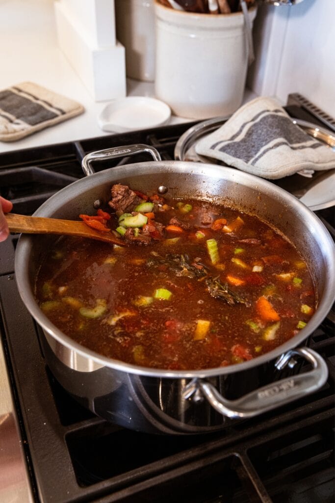 Hand scooping up pieces of carrot and celery in large metal stockpot filled with brown-colored soup with pieces of browned beef floating on the surface.