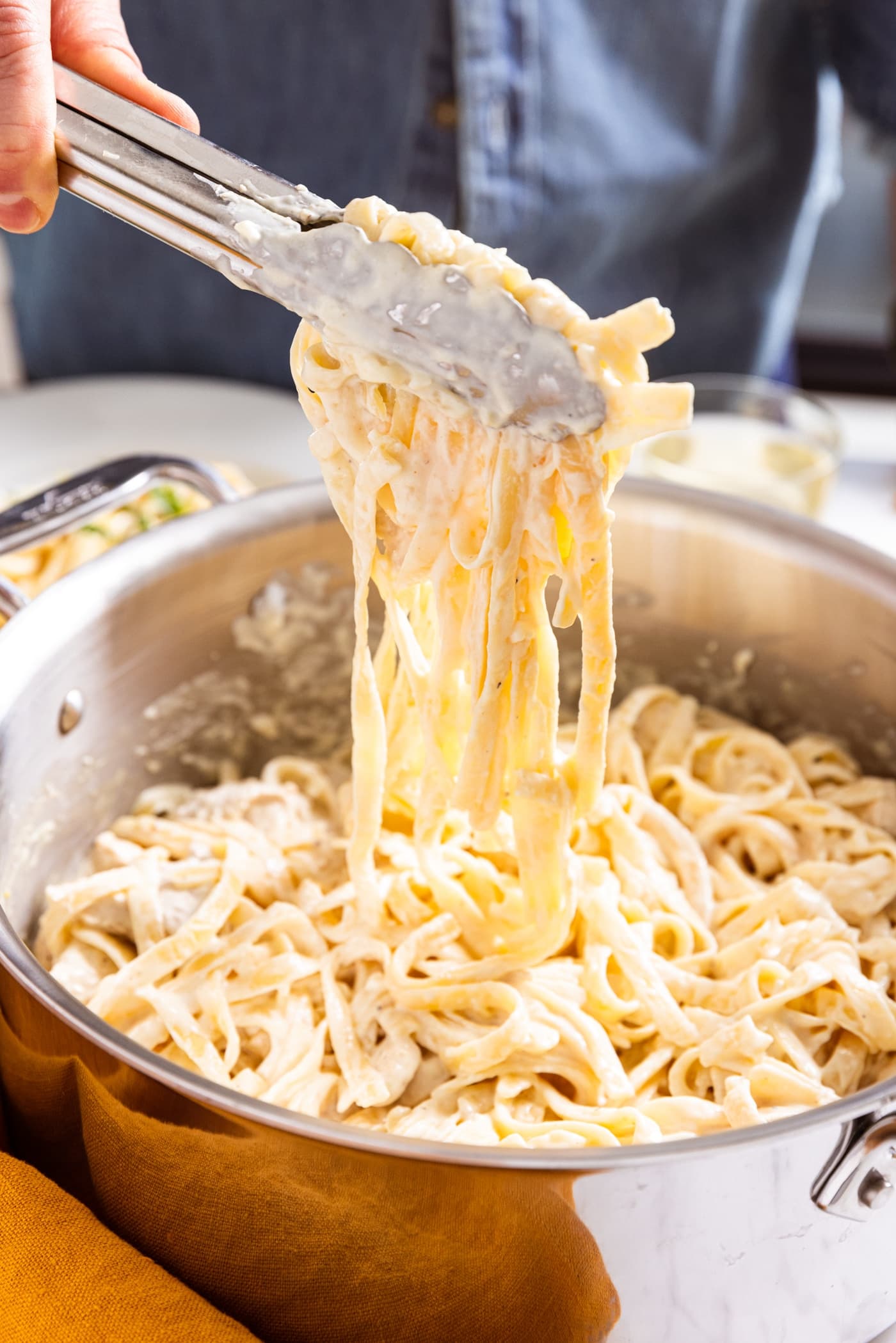 Tongs pulling up fettuccine with creamy alfredo sauce on noodles with pieces of chicken resting on pasta in large stockpot.
