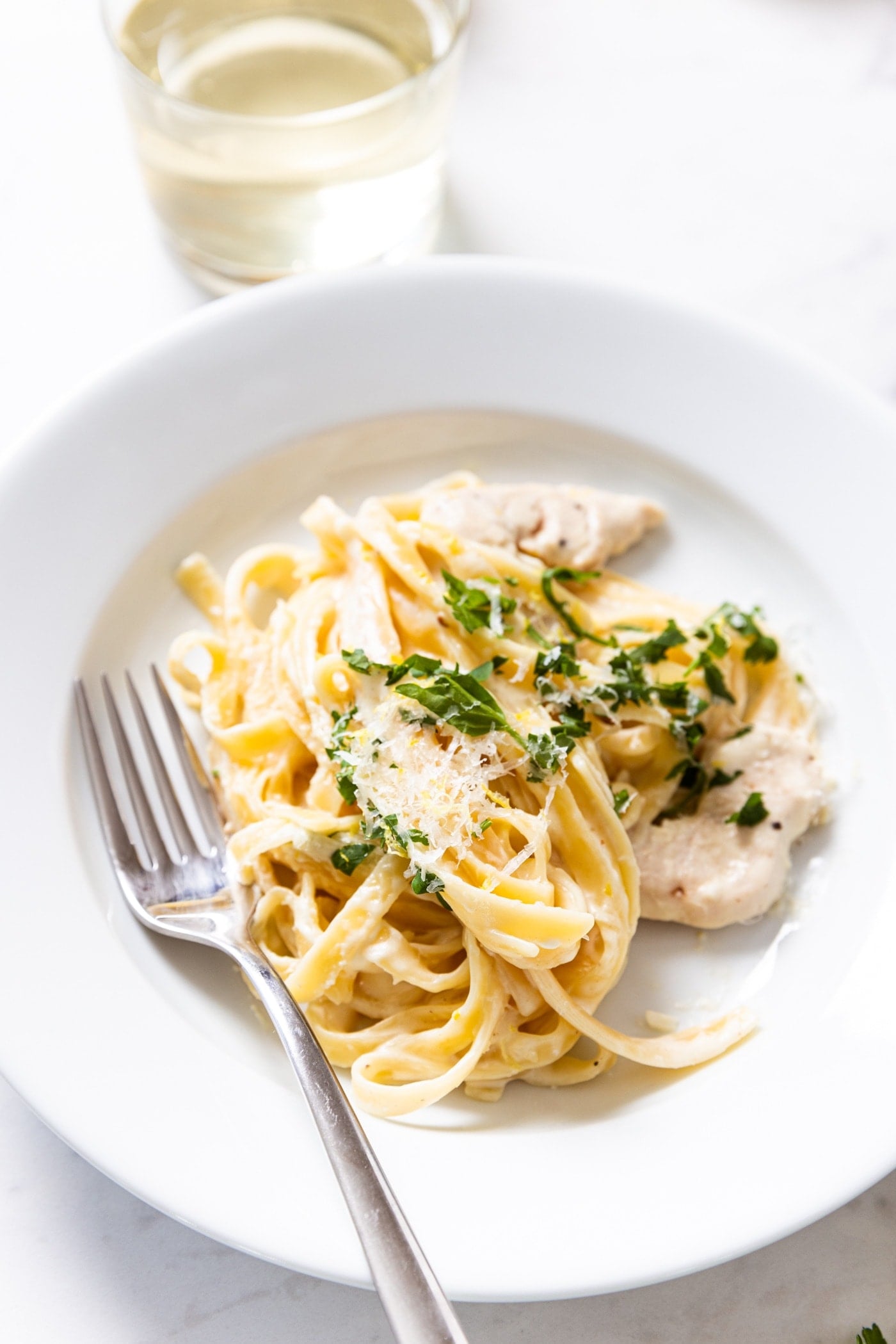 White plate filled with serving of chicken fettuccine alfredo topped with fresh parsley and Parmesan cheese with fork resting on side of plate with small glass on white wine in background.