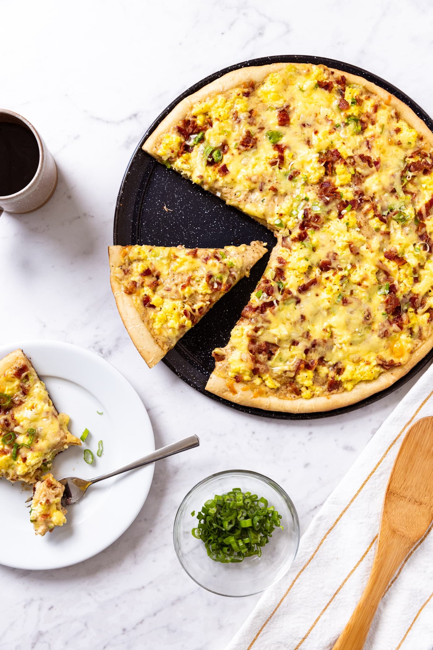 White marble surface with black pizza pan with bacon breakfast pizza after coming out of the oven with white plate with serving of pizza with small glass bowl filled with chopped scallion.