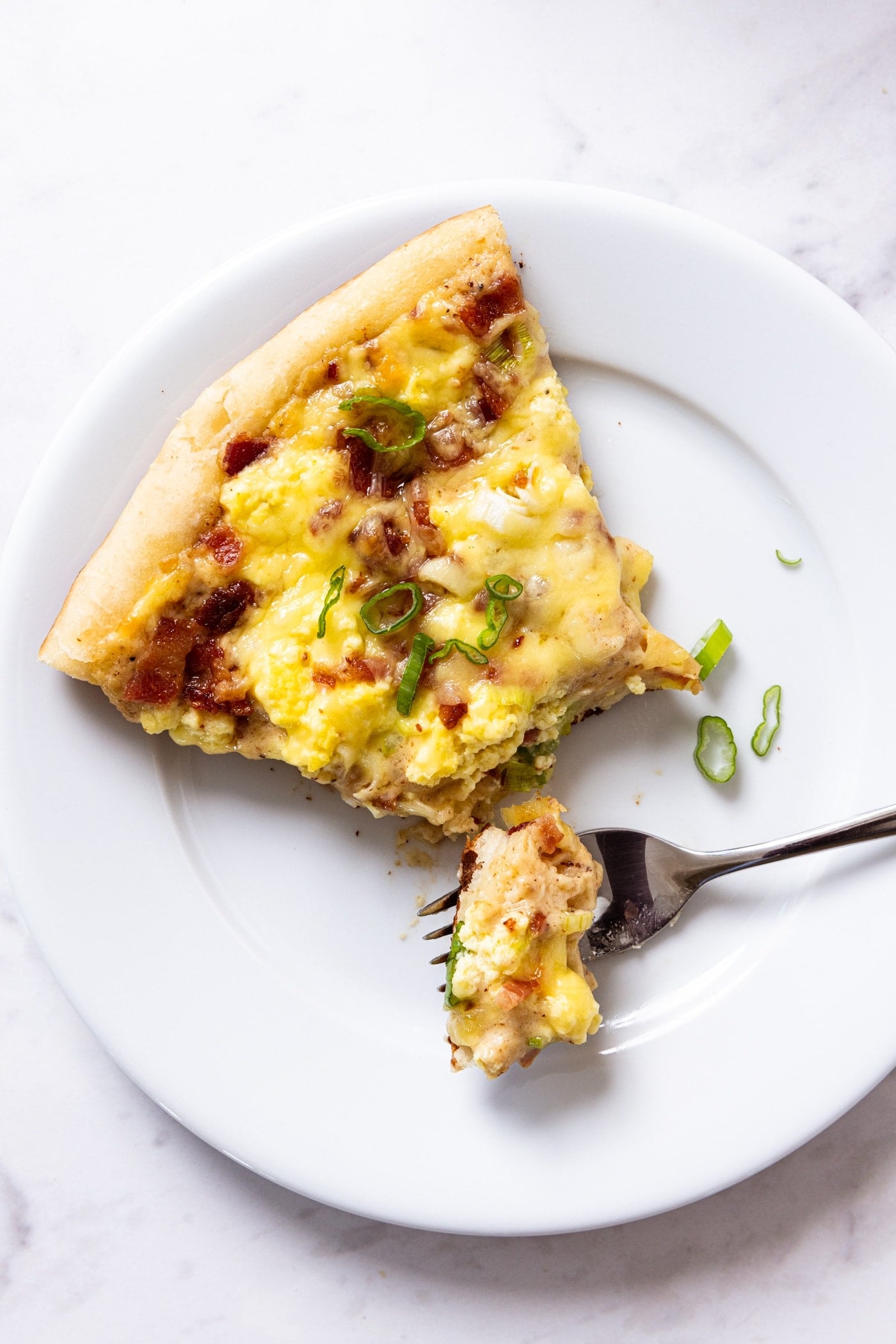 White plate with serving of bacon breakfast pizza with fork taking a bite from the slice with chopped scallion sprinkled over the top all on white marble surface.