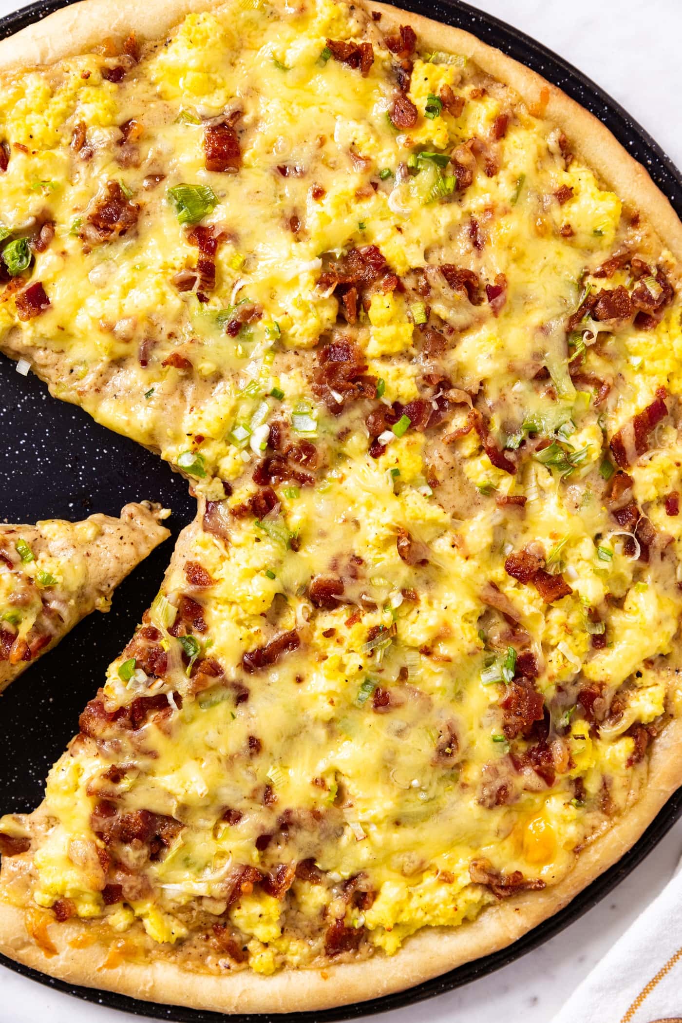 Black pizza pan filled with bacon breakfast pizza with a slice missing topped with bacon, eggs, cheese, and chopped scallion all on white marble surface.
