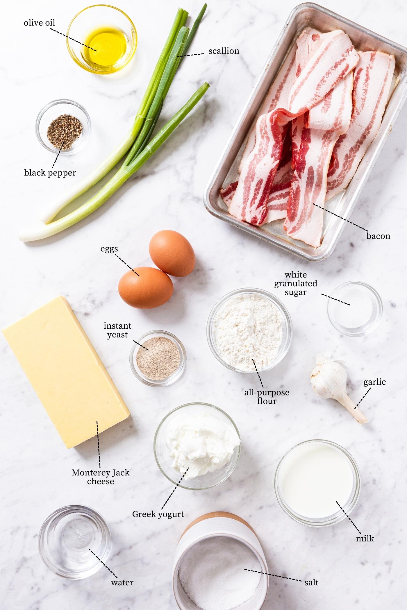 White marble surface filled with all ingredients needed to make bacon breakfast pizza including flour, eggs, scallion, bacon, cheese, black pepper, olive oil, and more.