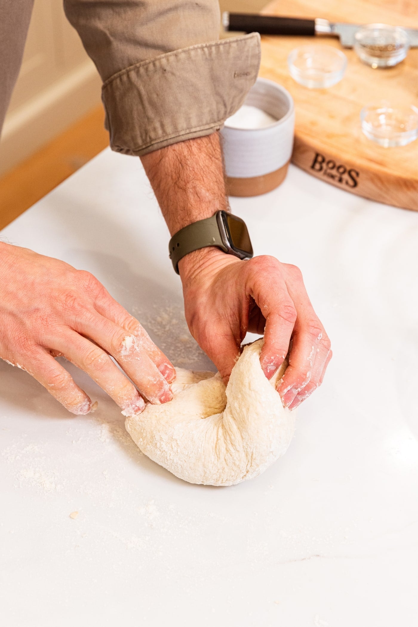 Two hands kneading dough on white surface with a lightly floured surface about to fold the dough over on itself.