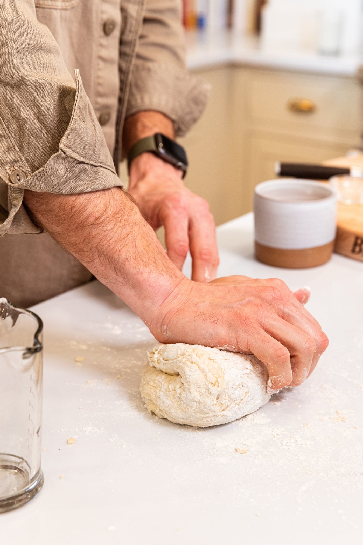 Hand kneading dough ball pressing it down in half after folding over on itself all on white surface.