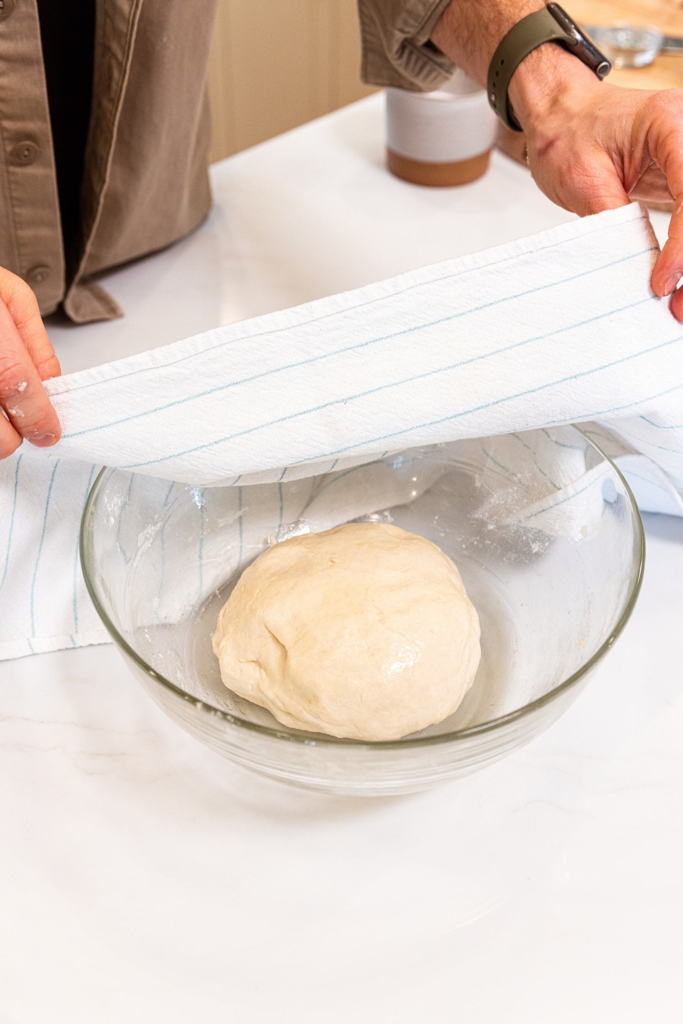 Hand placing white towel over the top of a glass bowl with dough ball oiled with olive oil all on white surface.