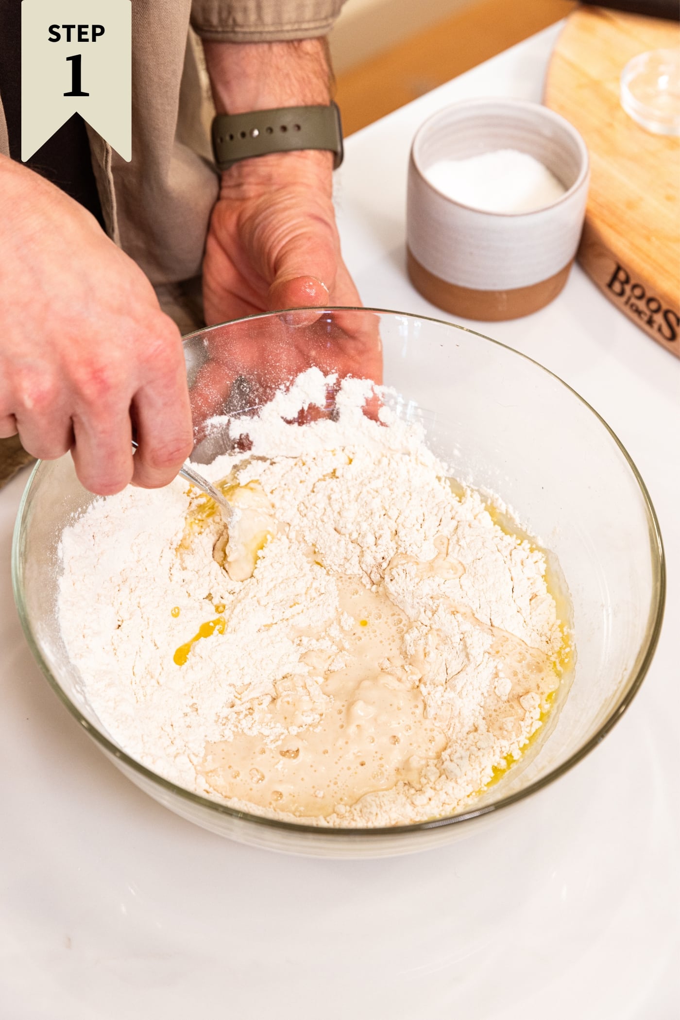 Glass bowl filled with flour and other ingredients needed to make a pizza dough with hand mixing with fork all on white surface.