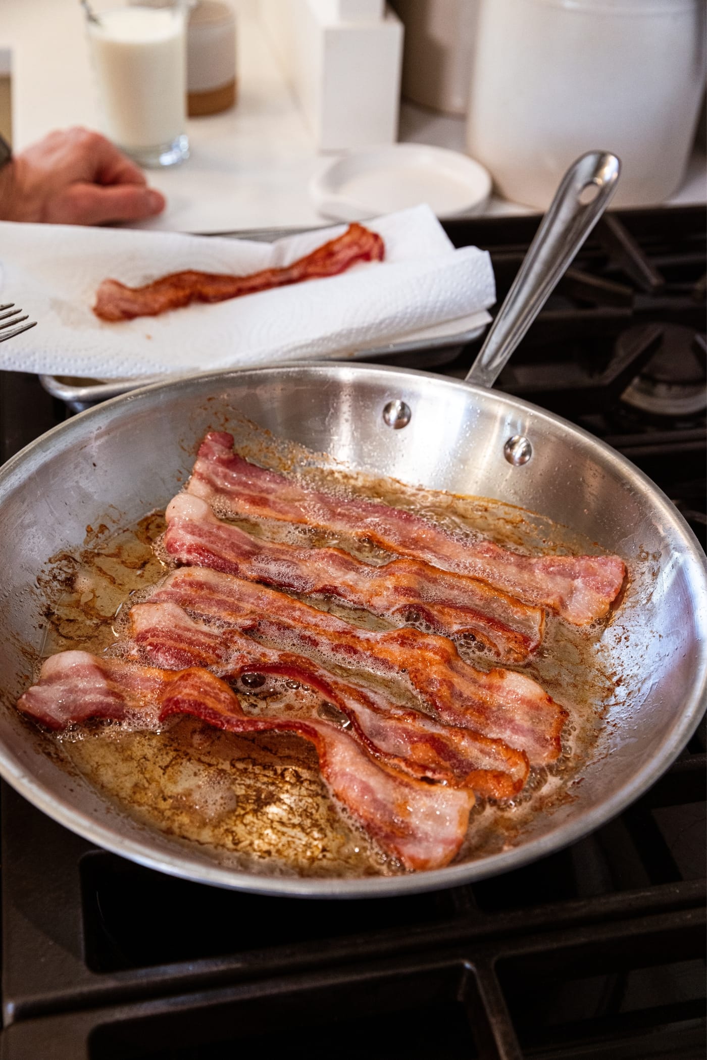 Cooking strips of bacon in metal skillet sitting on stovetop in bacon grease with piece of bacon sitting on paper towel in background.