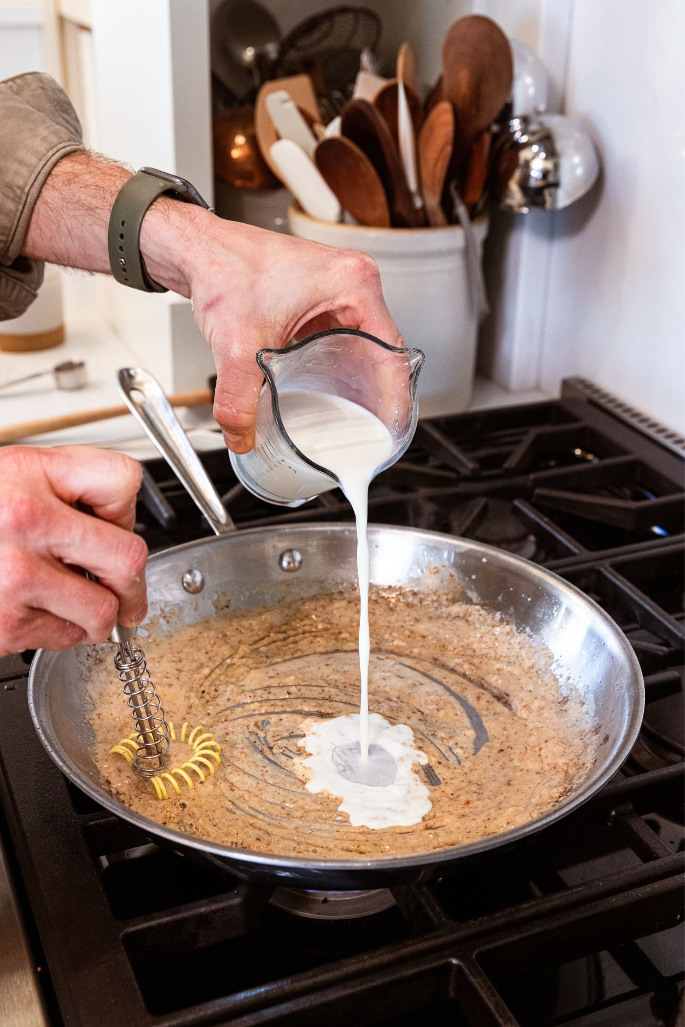 Hand holding glass measuring cup pouring milk into a metal skillet with browned bits and cooking flour and garlic on stovetop.