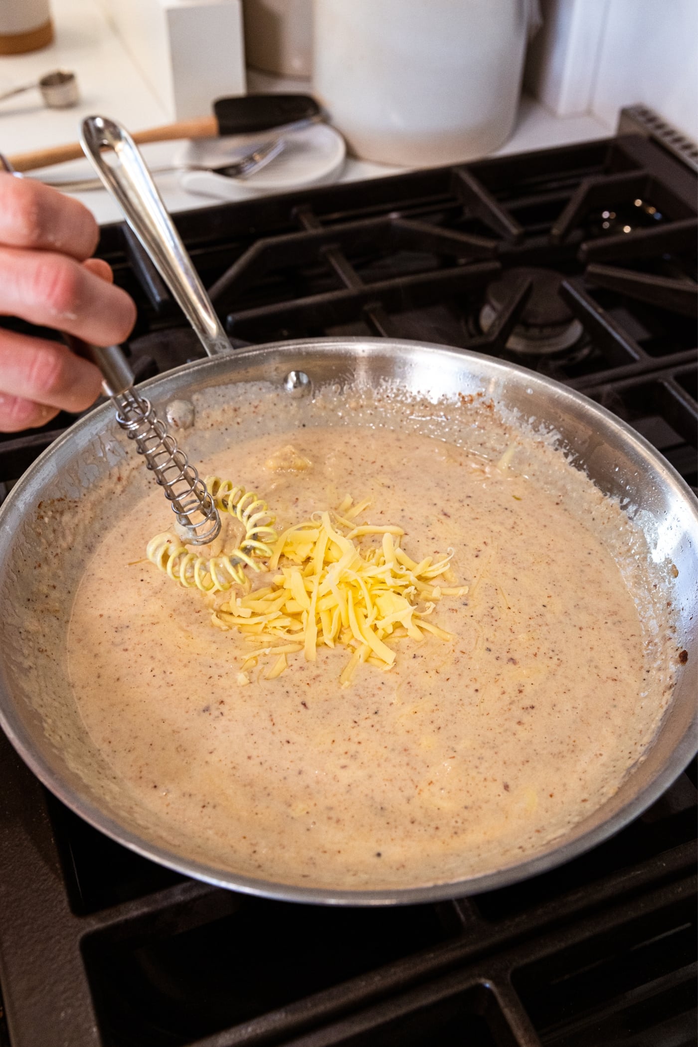 Shredded cheese resting on top of white sauce in metal skillet on stovetop about to be mixed togehter.
