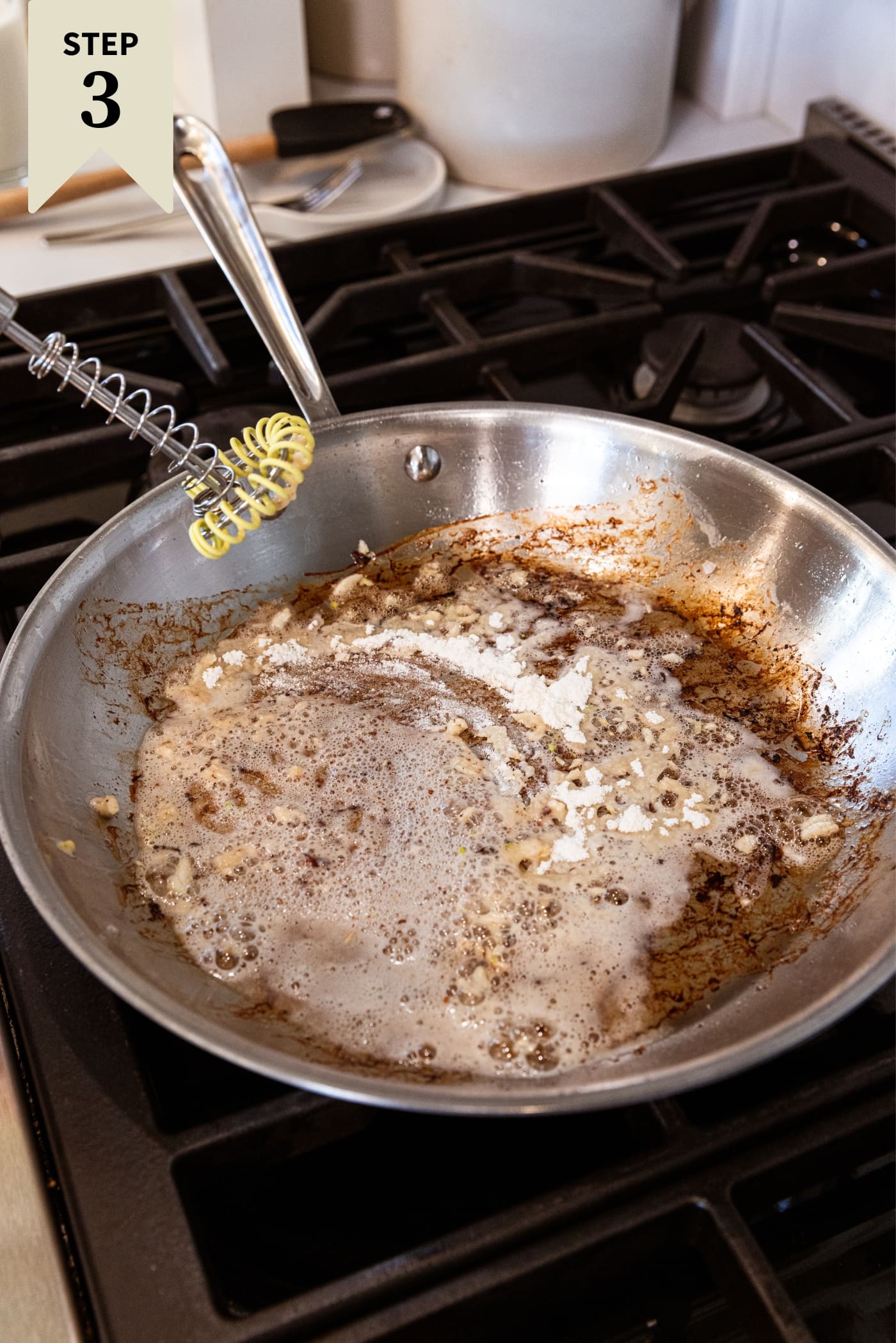 Metal skillet sitting on stovetop with browned bits on bottom of pan with garlic and flour about to be stirred together in pan all on stovetop.