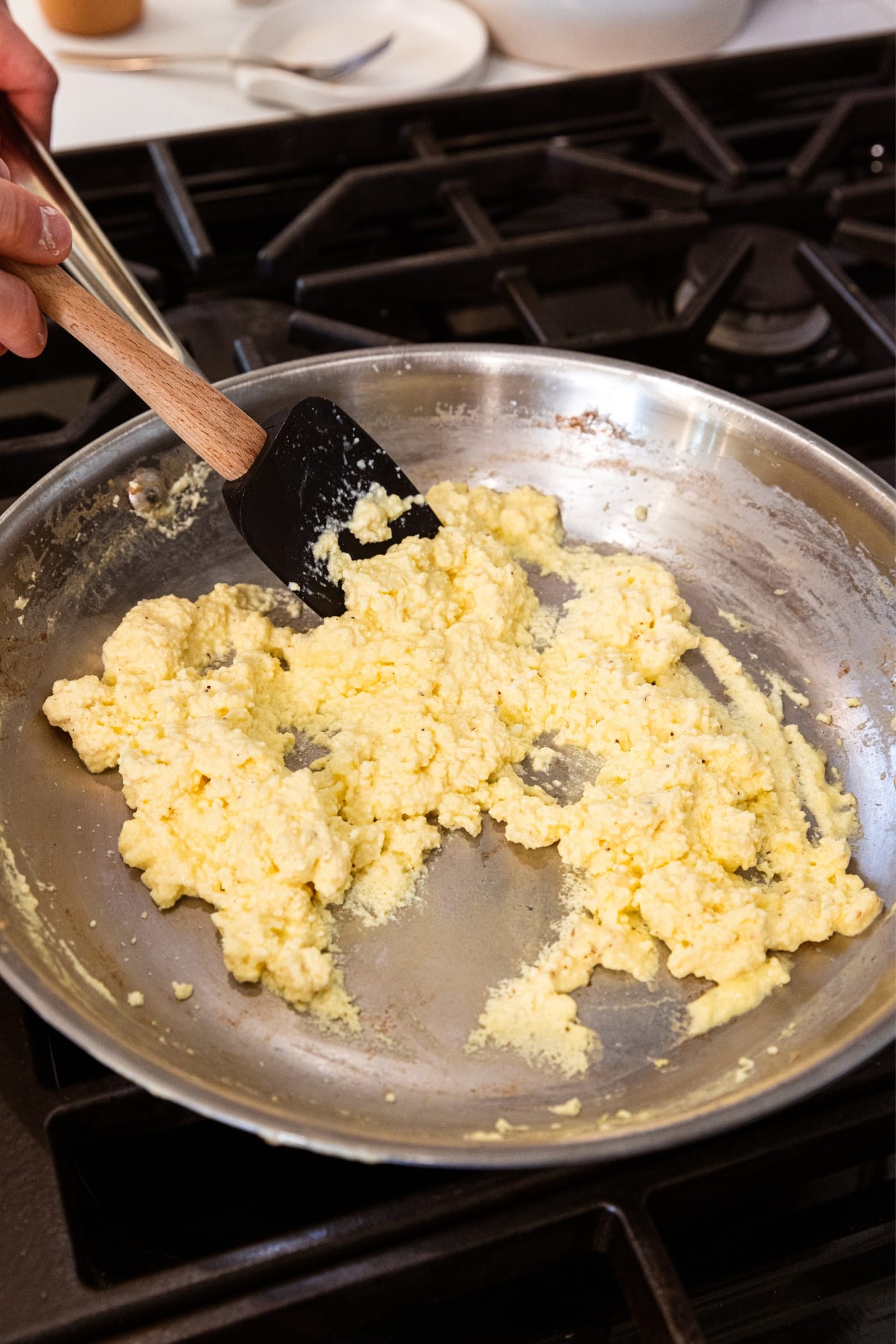 Hand using black spatula to mix together scrambled eggs in metal skillet on stovetop.