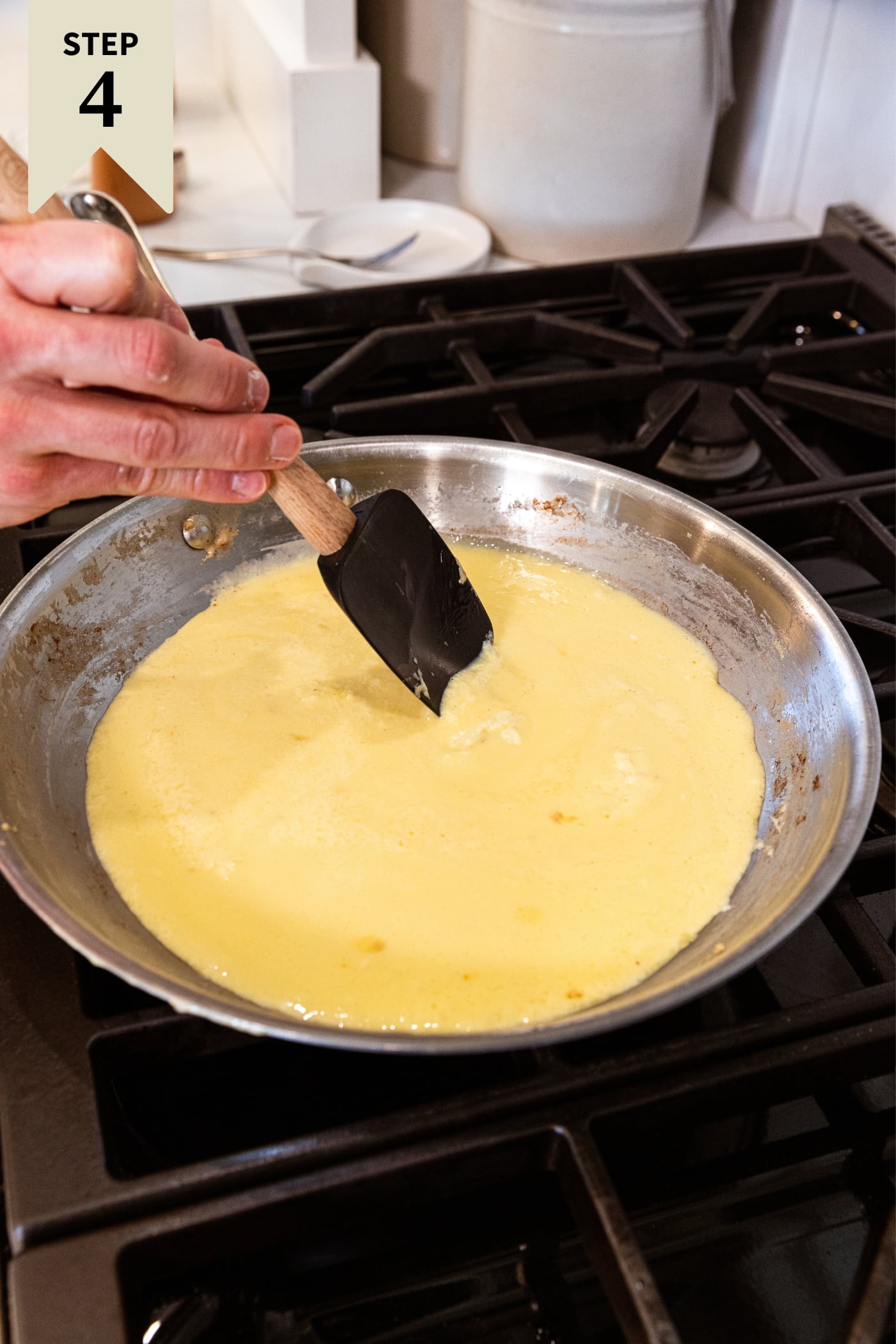 Hand holding black spatula stirring together egg mixture on metal skillet on stovetop before the eggs scramble.
