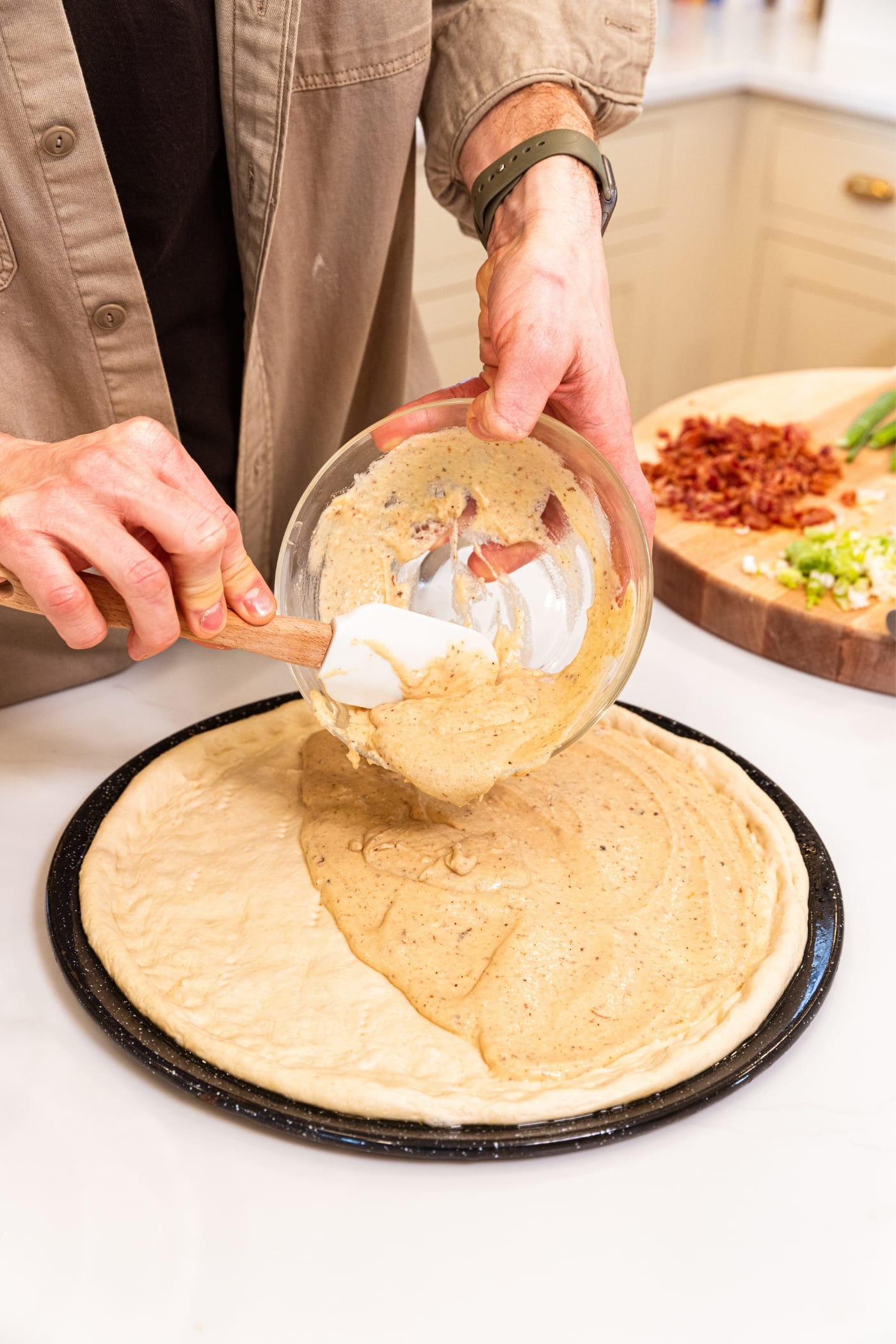 Hand holding glass bowl with cooled white cheese sauce pouring on top of formed pizza dough with white spatula.