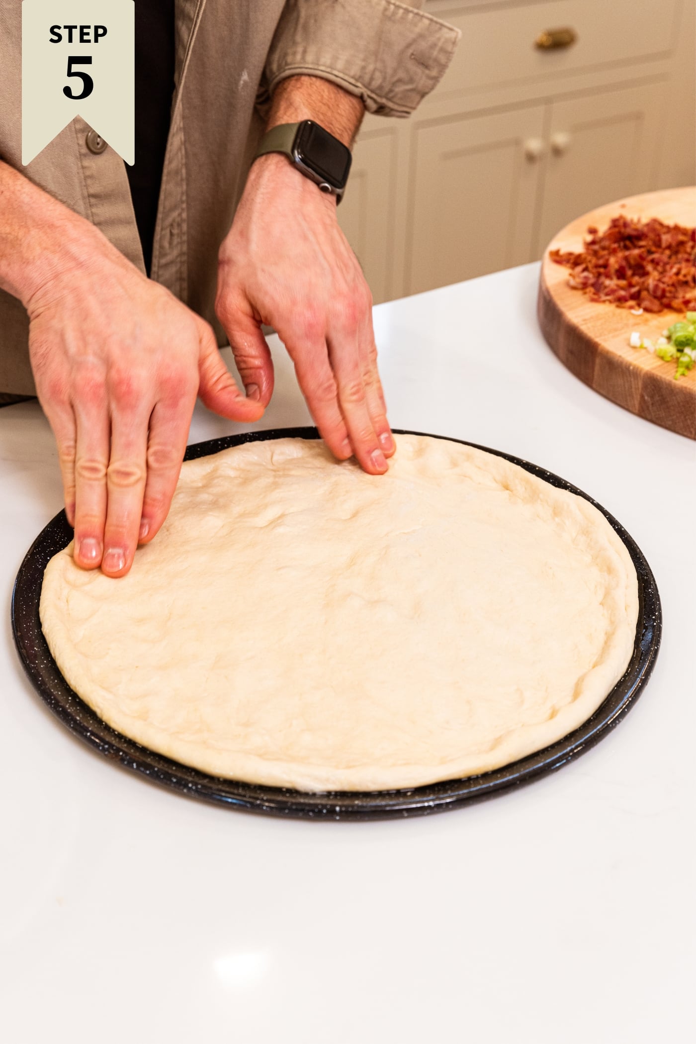 Two hands pressing out dough into a large circular pizza pan sitting on white surface with sliced toppings in background.