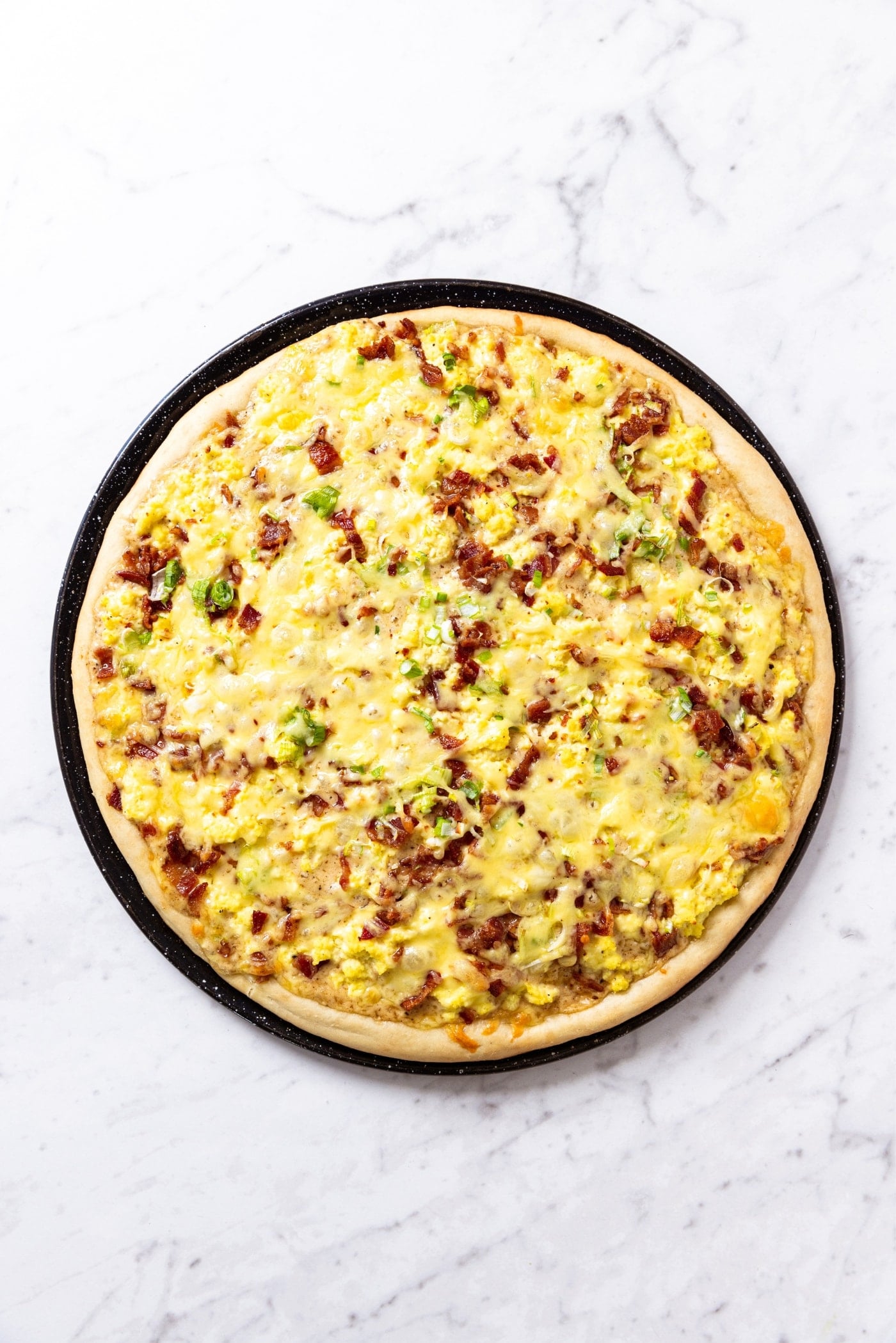 Black pizza pan sitting on marble surface with baked bacon breakfast pizza sitting on white marble surface.