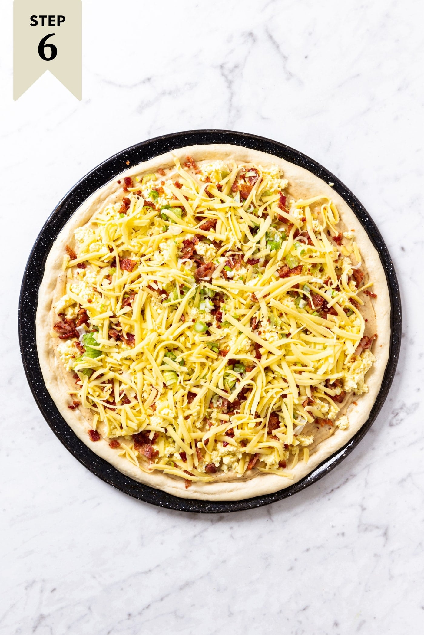 White marble surface with unbaked bacon breakfast pizza with shredded cheese on top sitting on black pizza pan.