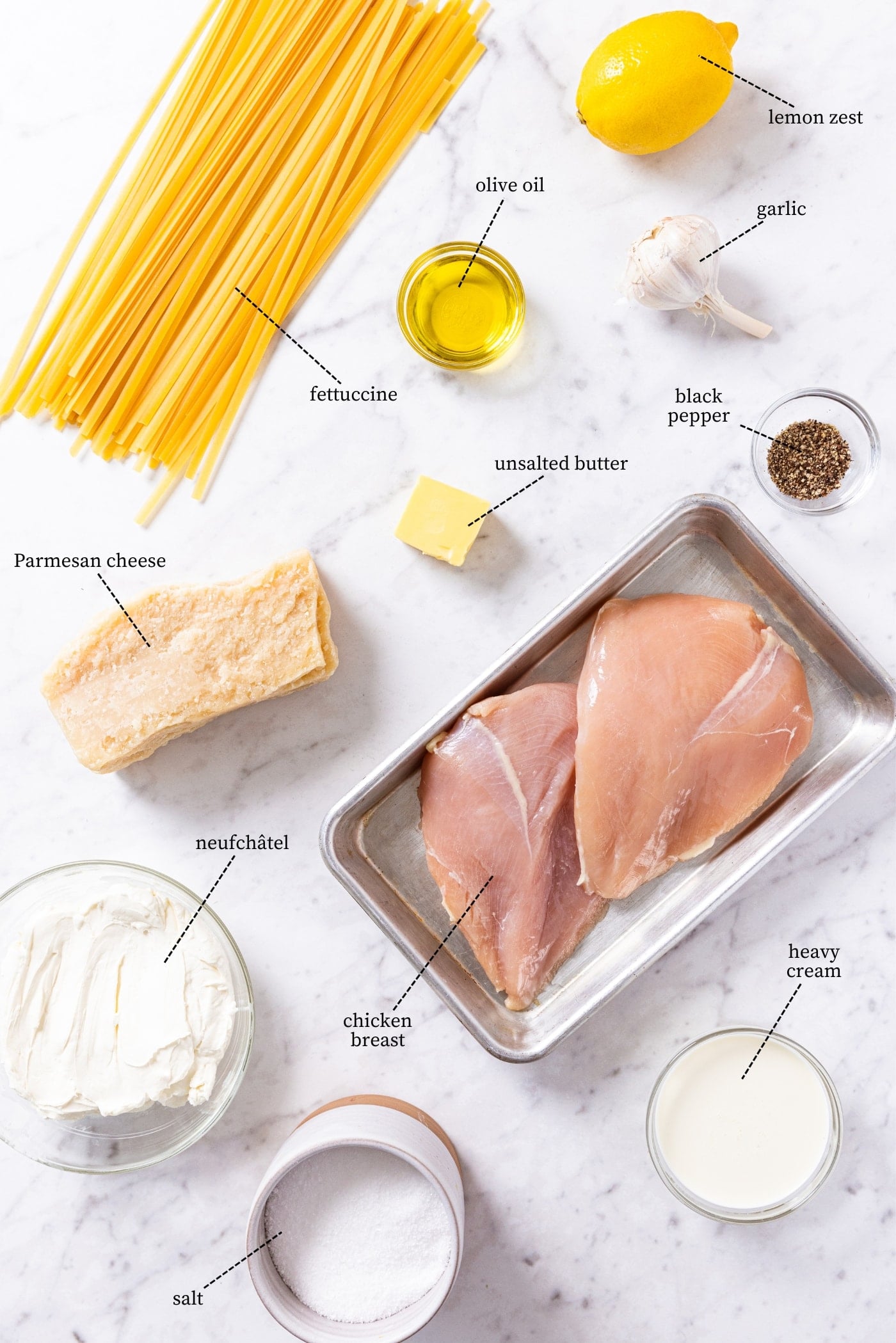 White marble surface filled with all ingredients needed to make chicken fettuccine alfredo including pasta, chicken breasts, lemon, olive oil, salt, heavy cream, and more.
