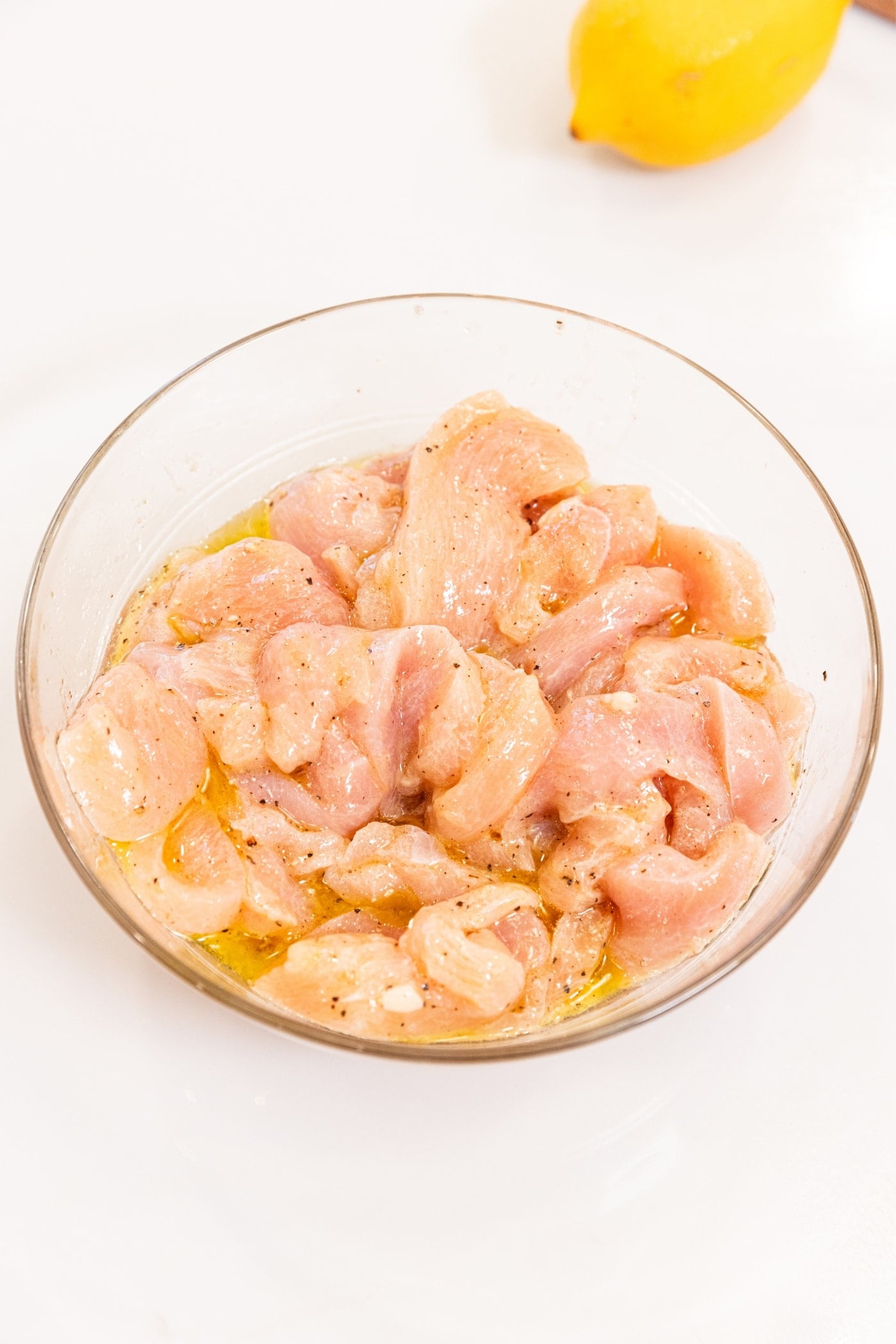 Glass bowl filled with raw pieces of chicken breast cutting into strips with olive oil and lemon zest mixed together all on white surface.
