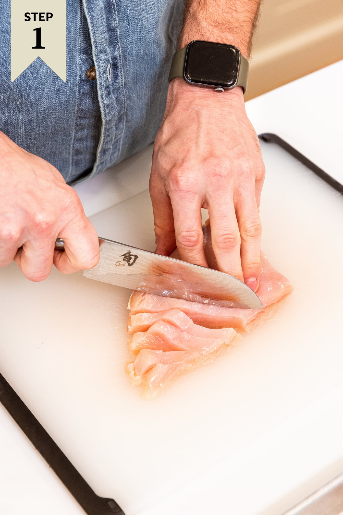 Hand using knife to slice raw chicken breasts into thin strips on white cutting board on kitchen countertop.