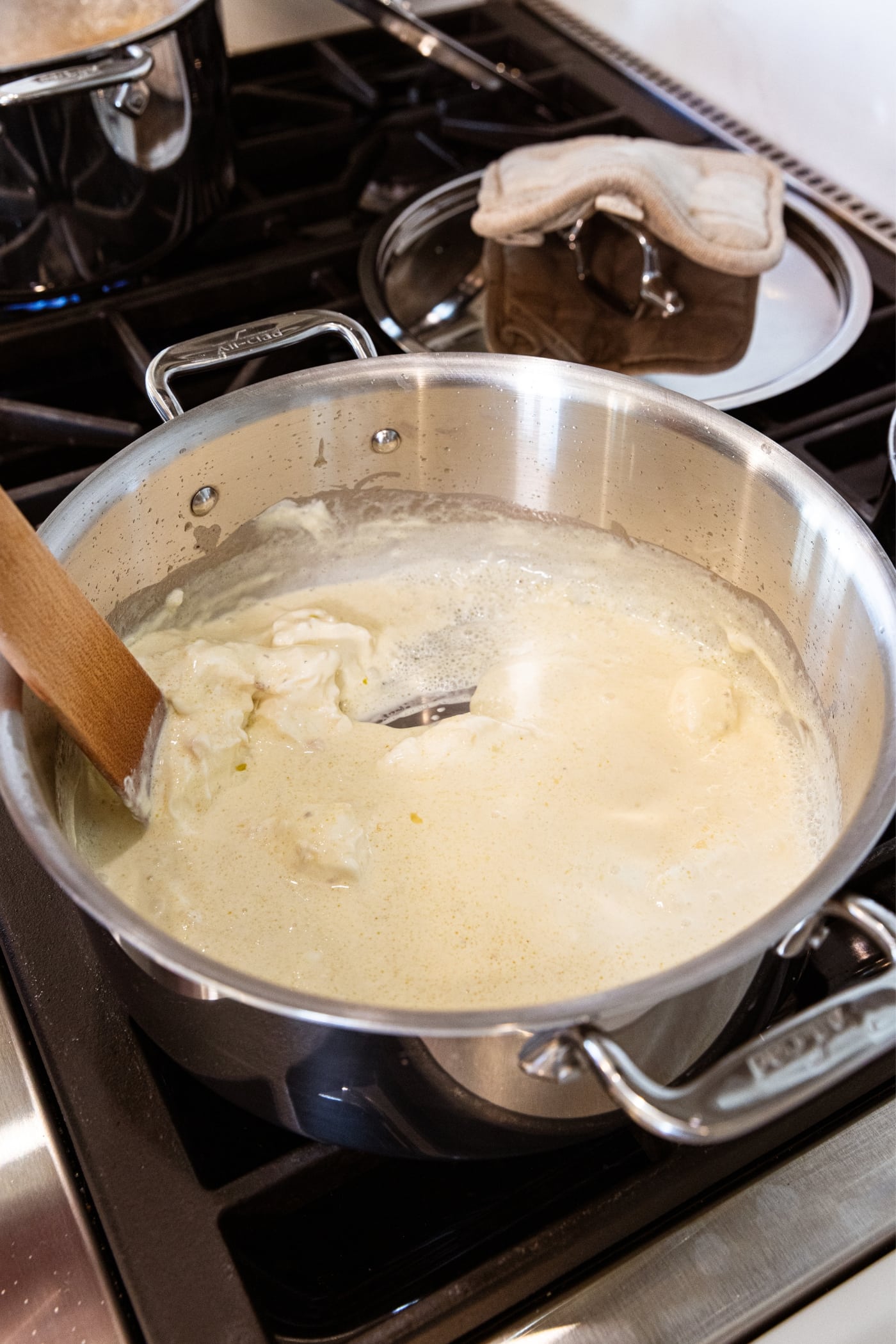 Creamy white colored sauce in metal stockpot with wooden spatula pushing mixture around showing thickness of the sauce.