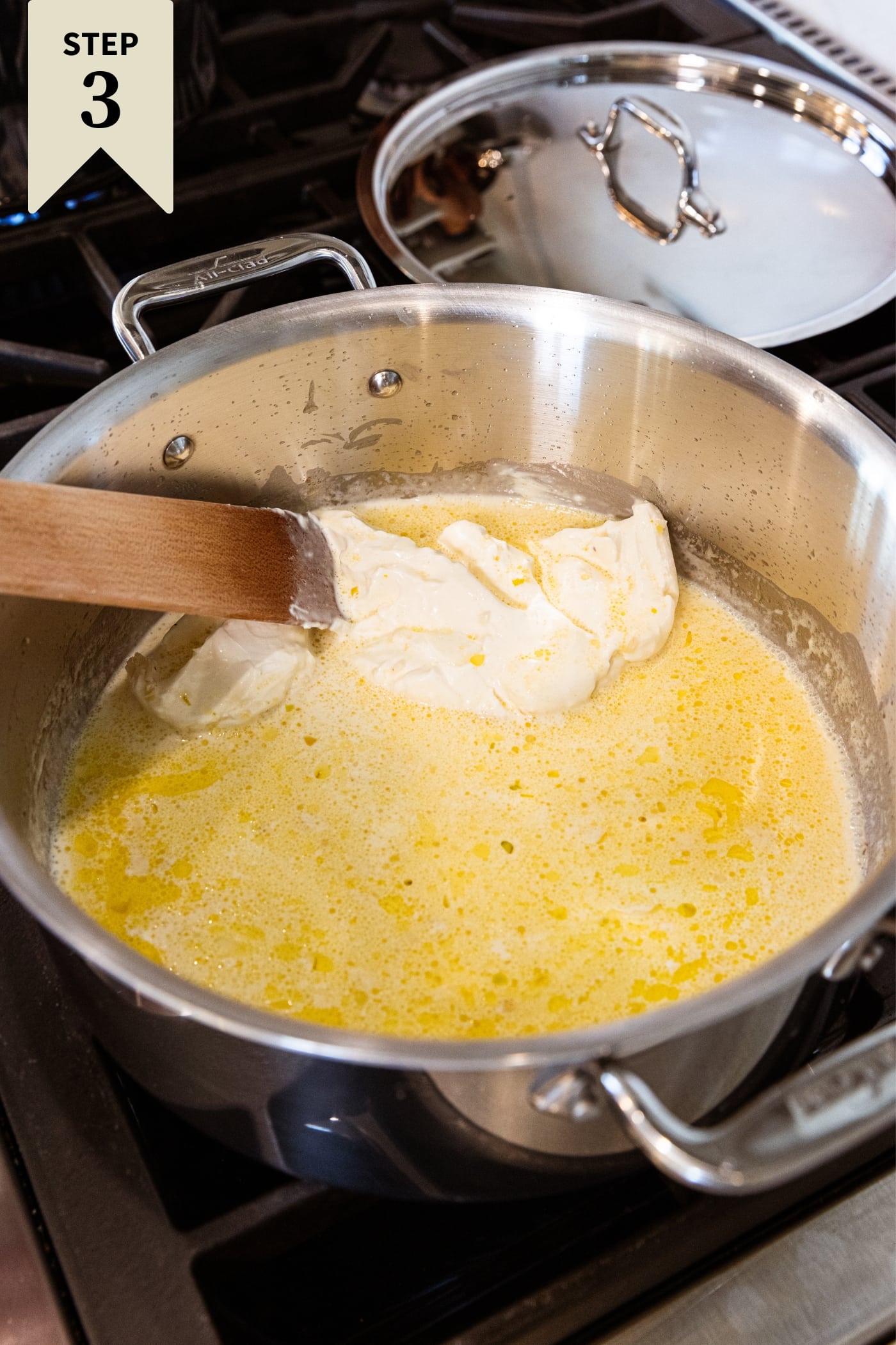 Large metal stockpot on stovetop with melting heavy cream and cream cheese over heat with wooden spatula mixing everything together slowly.