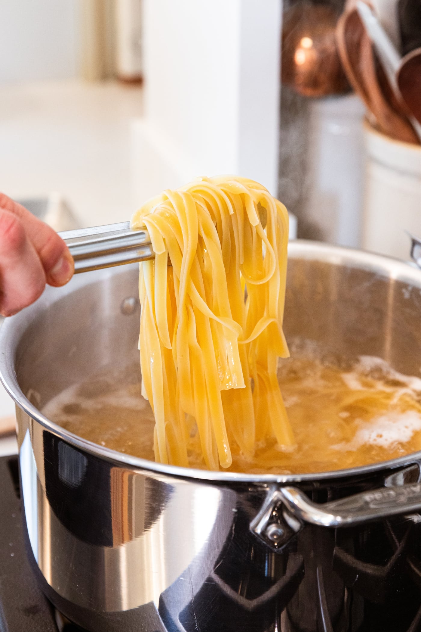 Hand using tongs to light cooked fettuccine noodles out of a large metal stockpot over high meat on stovetop.