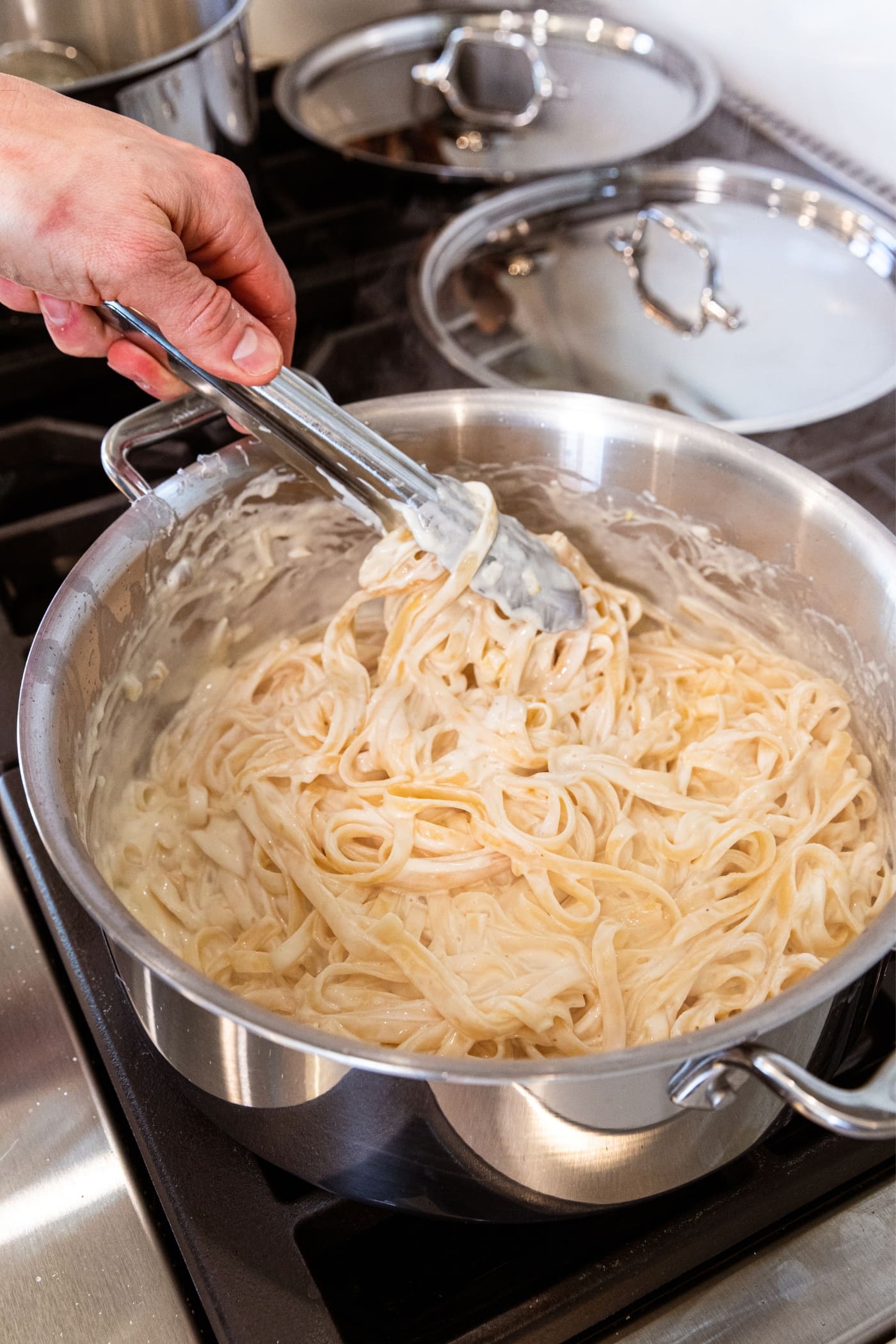 Hand using tongs to mix together fettuccine noodles with alfredo sauce in large stockpot on stovetop.