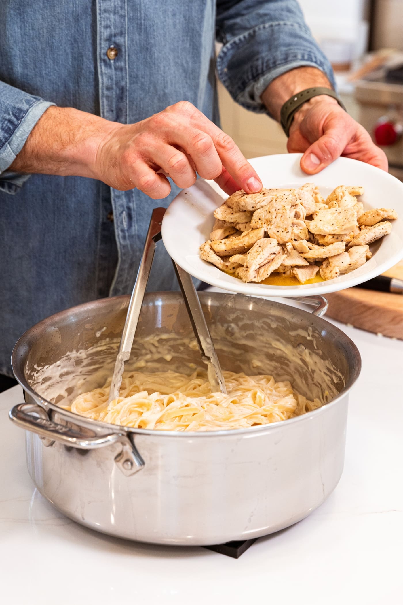 Hand holding white plate about to pour cooked chicken pieces into alfredo sauce with fettuccine noodles sitting on white surface.