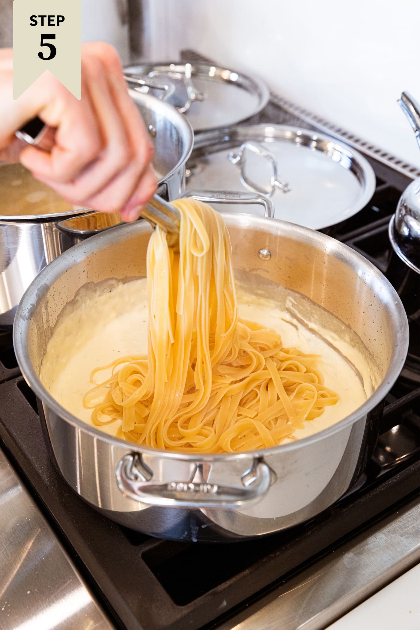 Hand using tongs to place cooked fettuccine noodles in large stockpot filled with simmering alfredo sauce on stovetop.