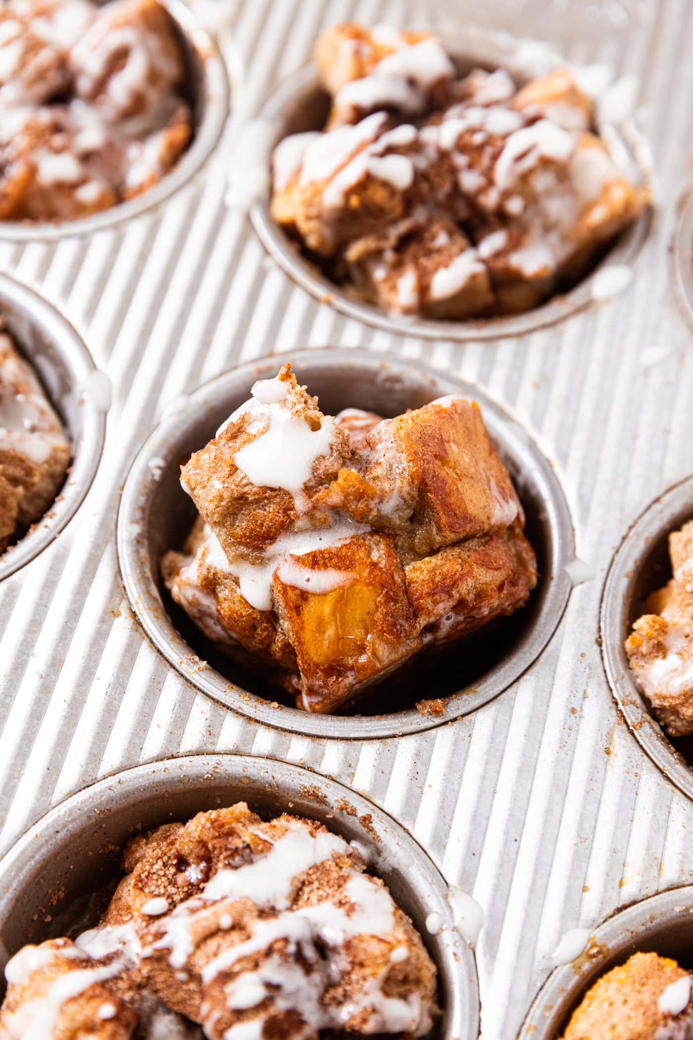 Gray muffin pan with French toast muffins resting in pan drizzled with thin white sugar glaze on top with one muffin set on its side to see the interior texture.