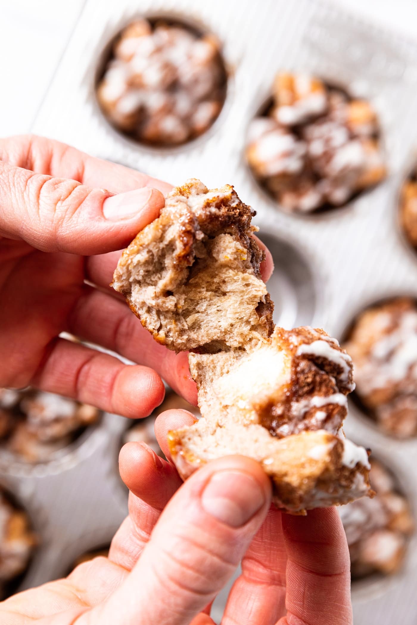 Two hands tearing French toast muffin in half showing the interior texture made up on cubed bread and cinnamon flecks with rest of muffins below.