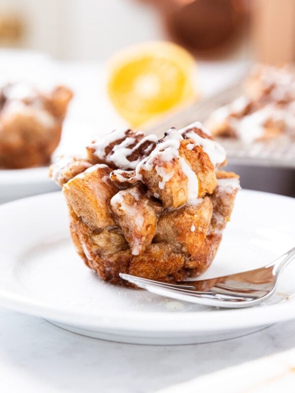 White plate with French toast muffin drizzled with thin white glaze sitting before being eaten with fork also resting on the plate with sliced orange and rest of muffins in background.