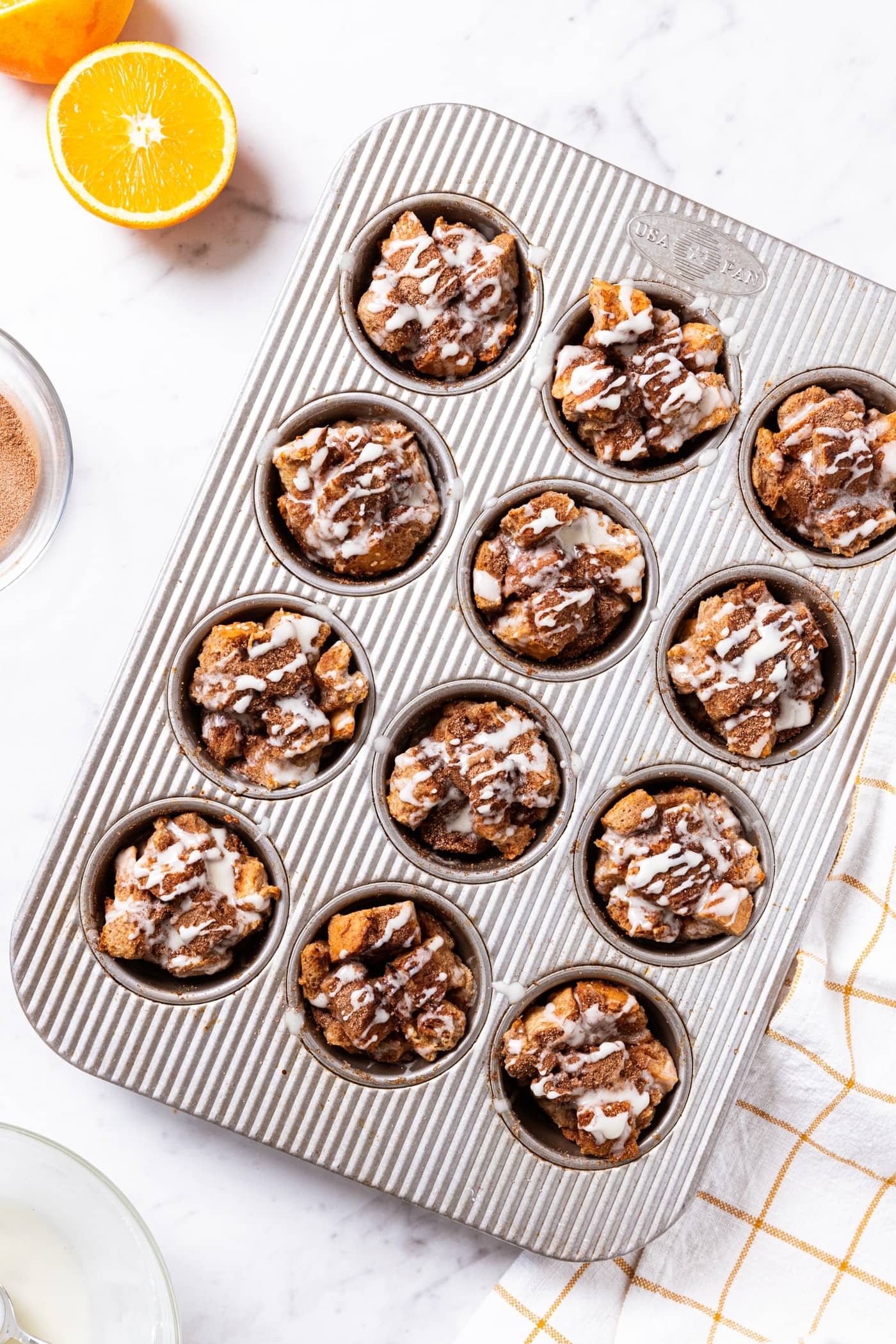 Gray muffin pan filled with baked French toast muffins sitting on white marble surface with thin white glaze spread over the top of each muffin with orange resting to the side.