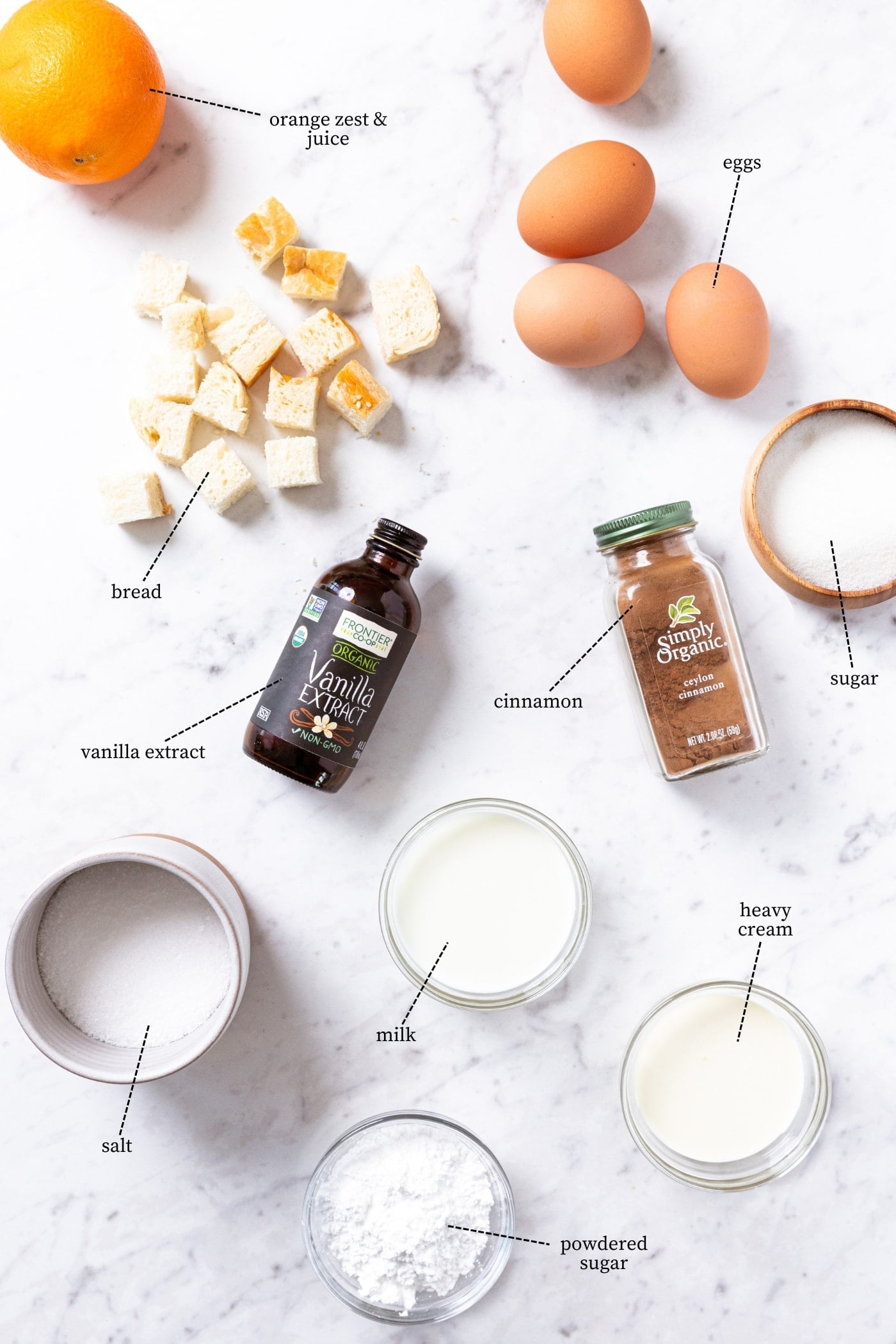 White marble surface filled with all ingredients needed to bake French toast muffins including eggs, orange, vanilla, bread cubes, cinnamon, sugar, cream, and more.