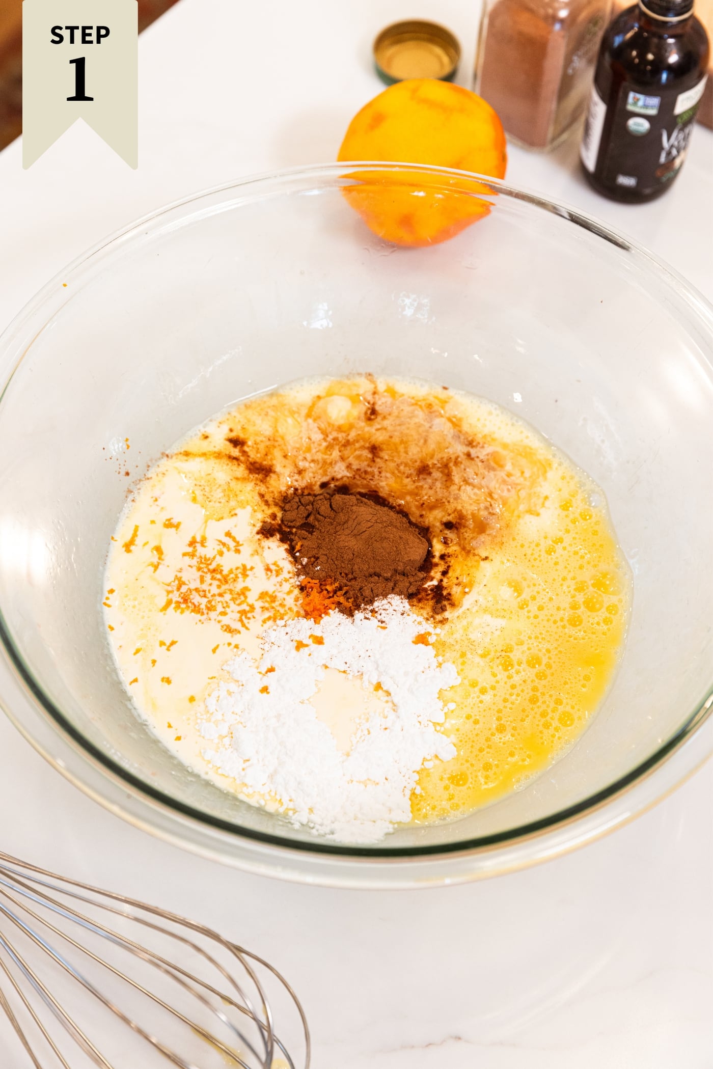 Glass bowl filled with ingredients needed to make French toast muffins including cinnamon, eggs, flour, and sugar all on white surface.