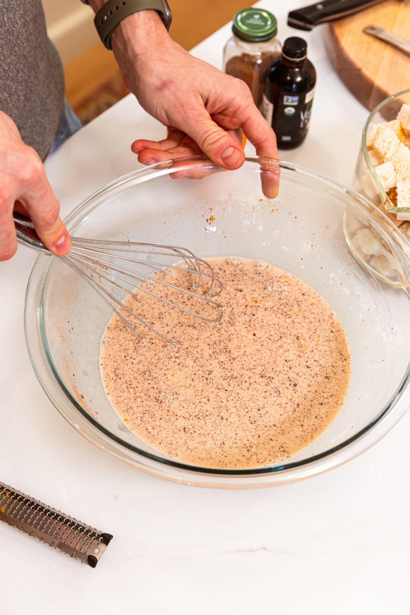Hand using metal whisk to whisk up the ingredients for French toast muffin custard base in large glass bowl all on white surface.
