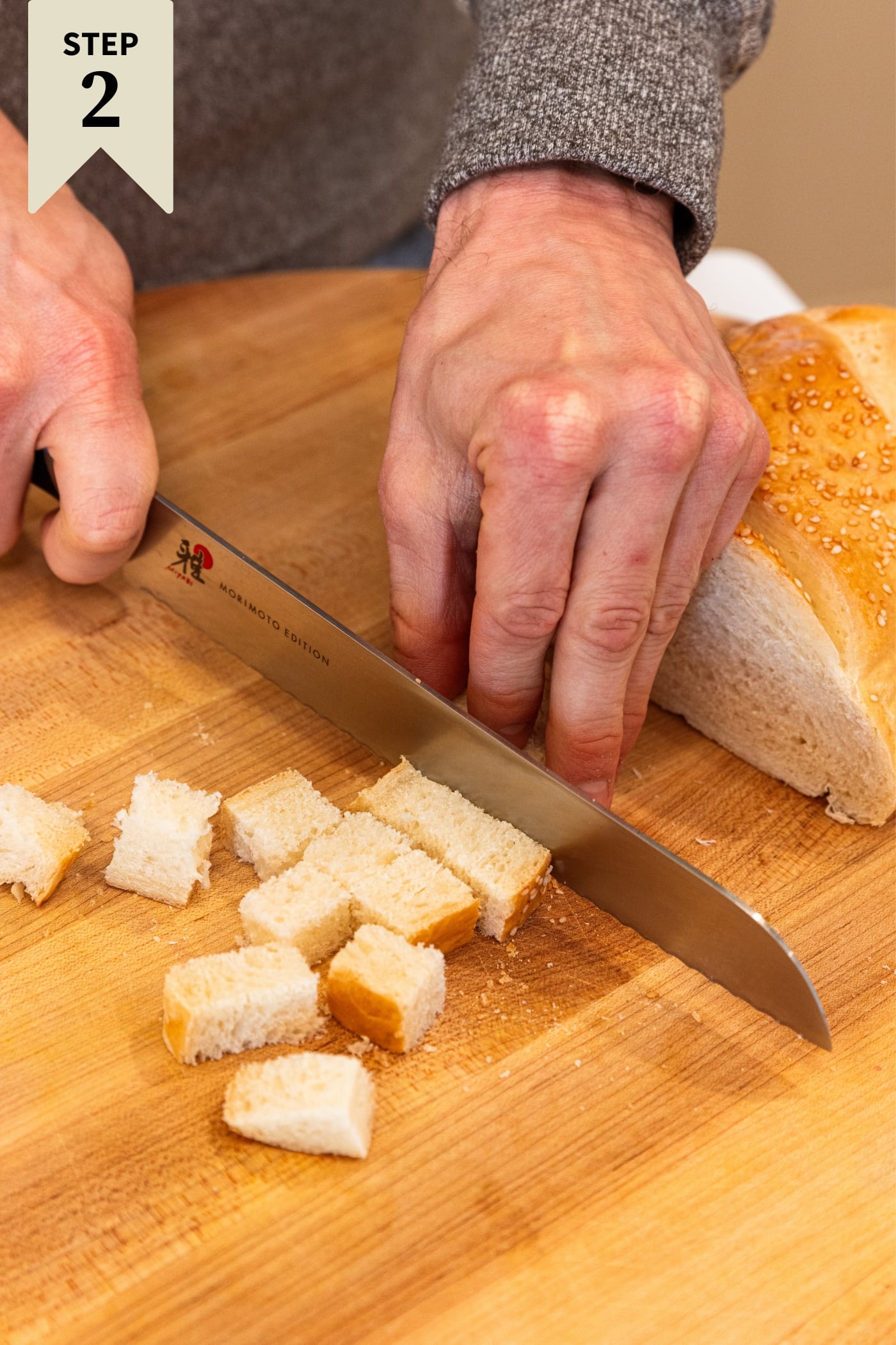 Hand using knife to slice up French bread into chunks of small cubes on wood cutting board.