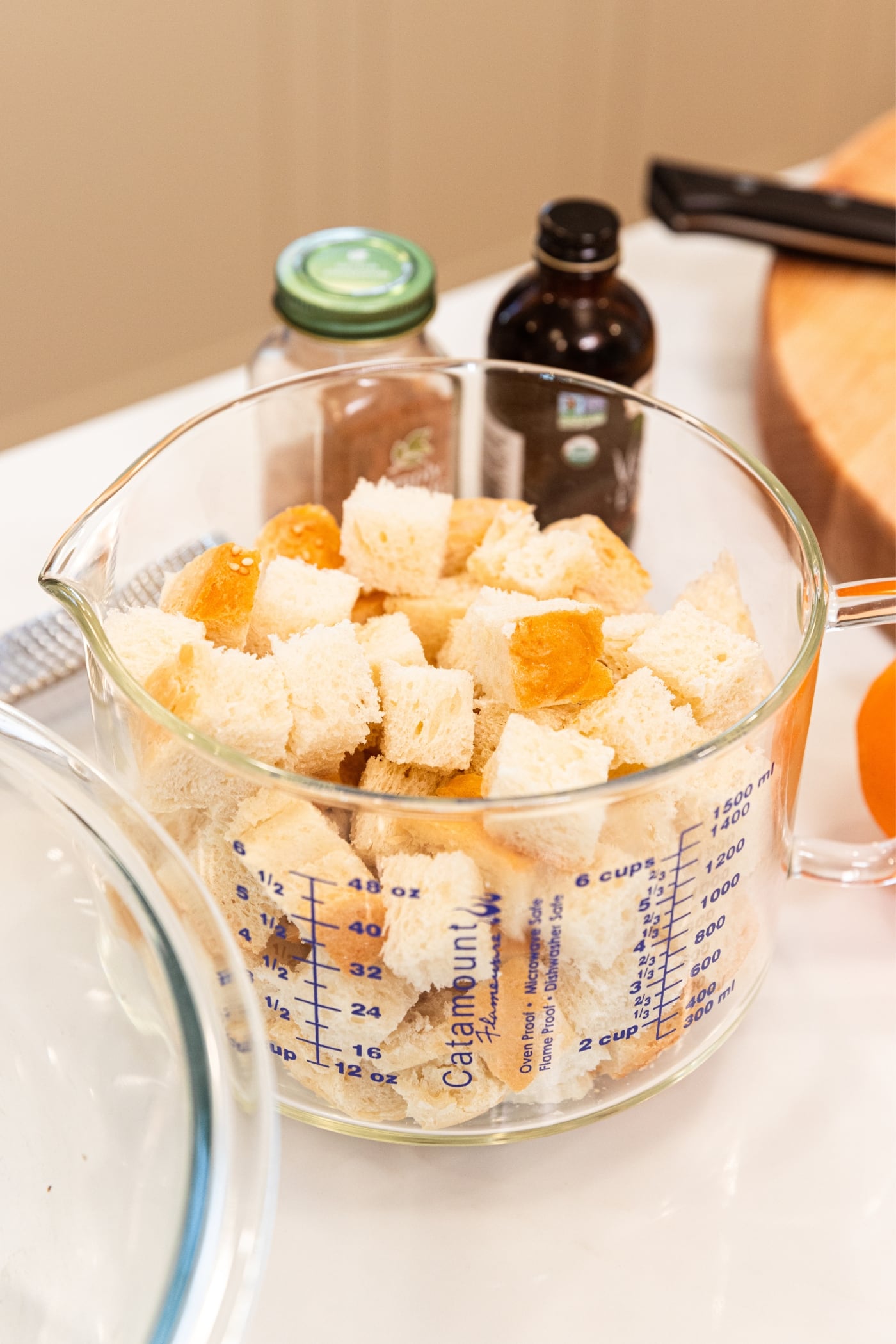 Glass measuring container filled with cubed chunks of French bread sitting on white countertop with spices and vanilla extract in background.