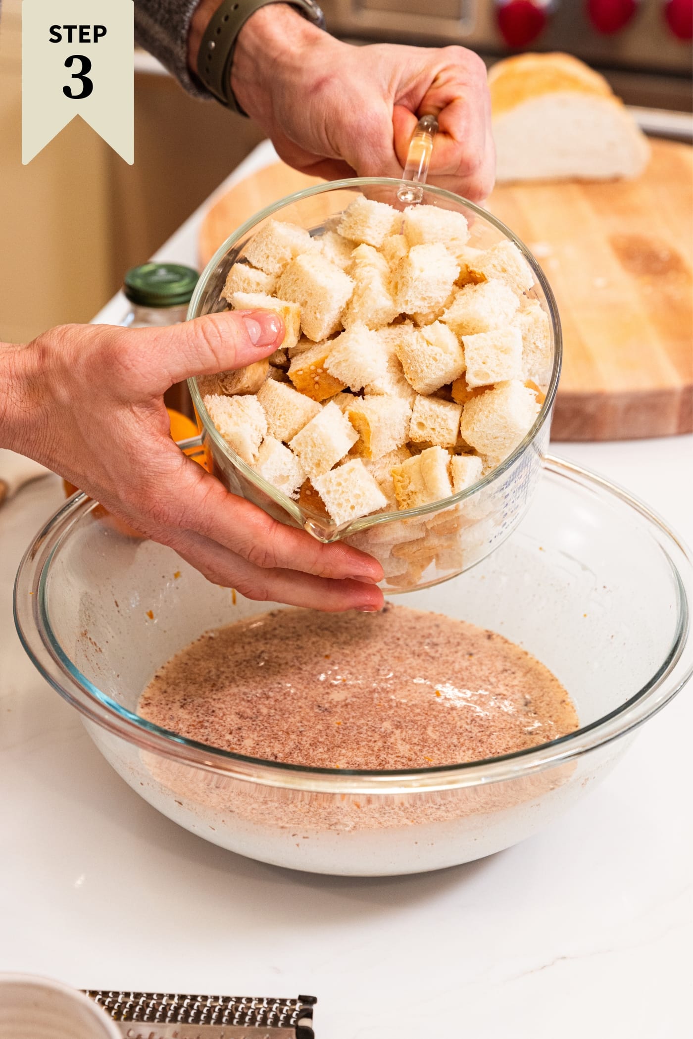 Glass measuring cup filled with cubed bread being poured into large glass bowl filled with custard base with cinnamon all sitting on white countertop.