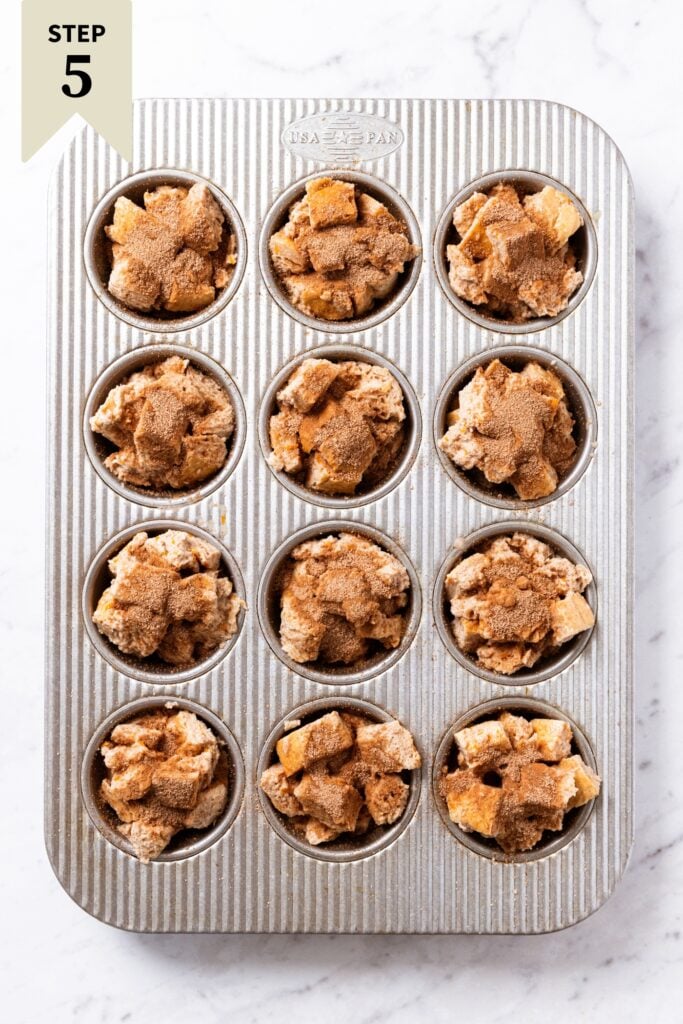 Gray muffin pan sitting on white marble surface filled with unbaked muffins filling each muffin tin with cinnamon sugar mixture sprinkled over the top.