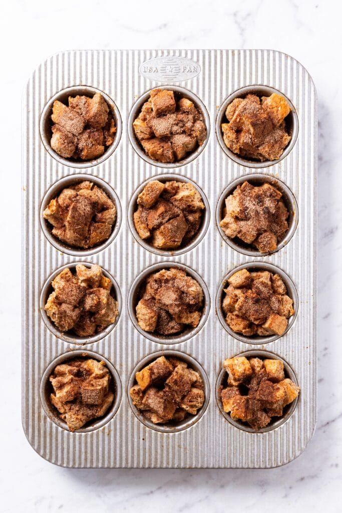 Gray muffin pan filled with baked French toast muffins sitting on white marble surface right after coming out of the oven.