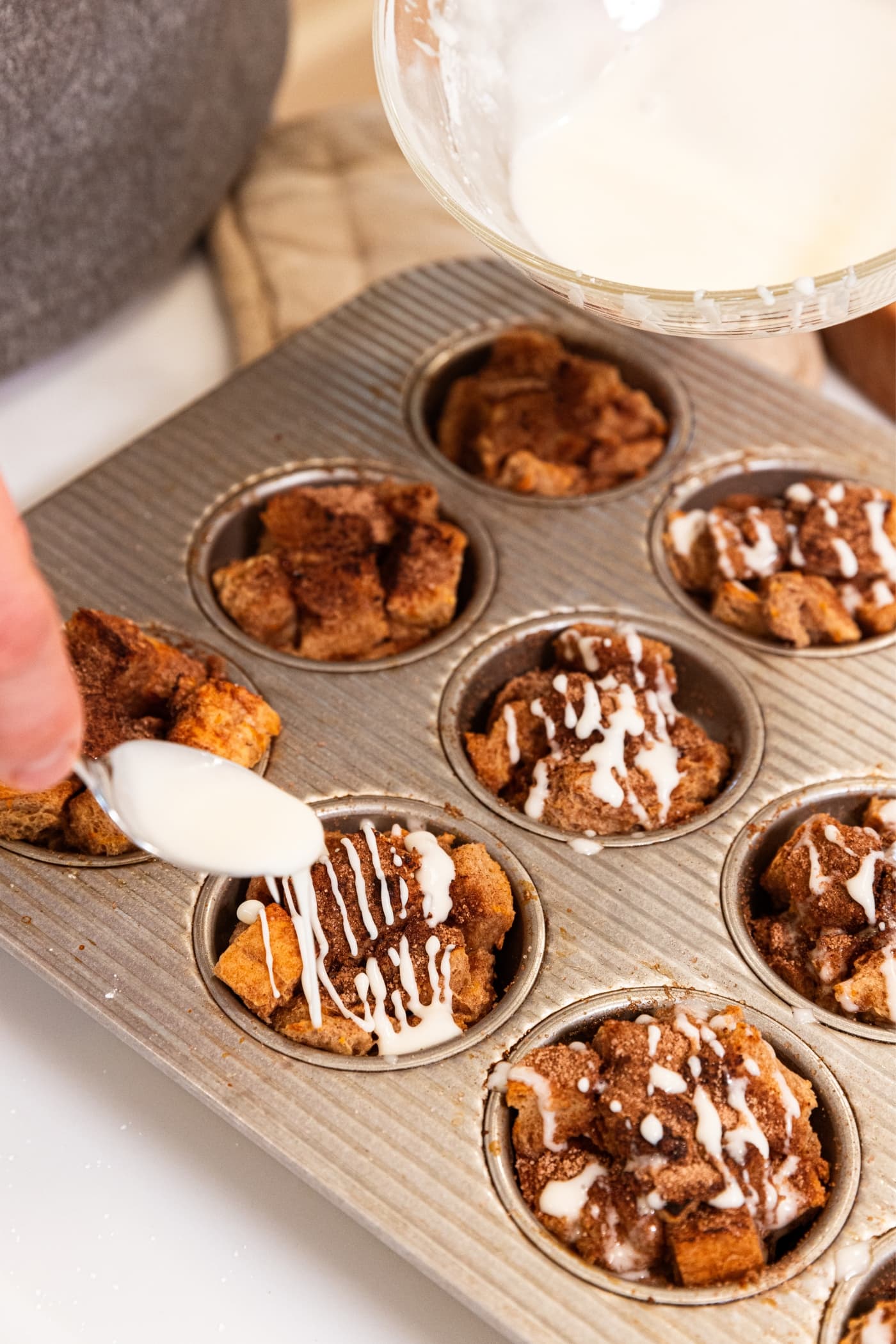 Hand using a white spatula to drizzle thin white sugar glaze over the top of French toast muffins still sitting in the muffin pan after being baked.