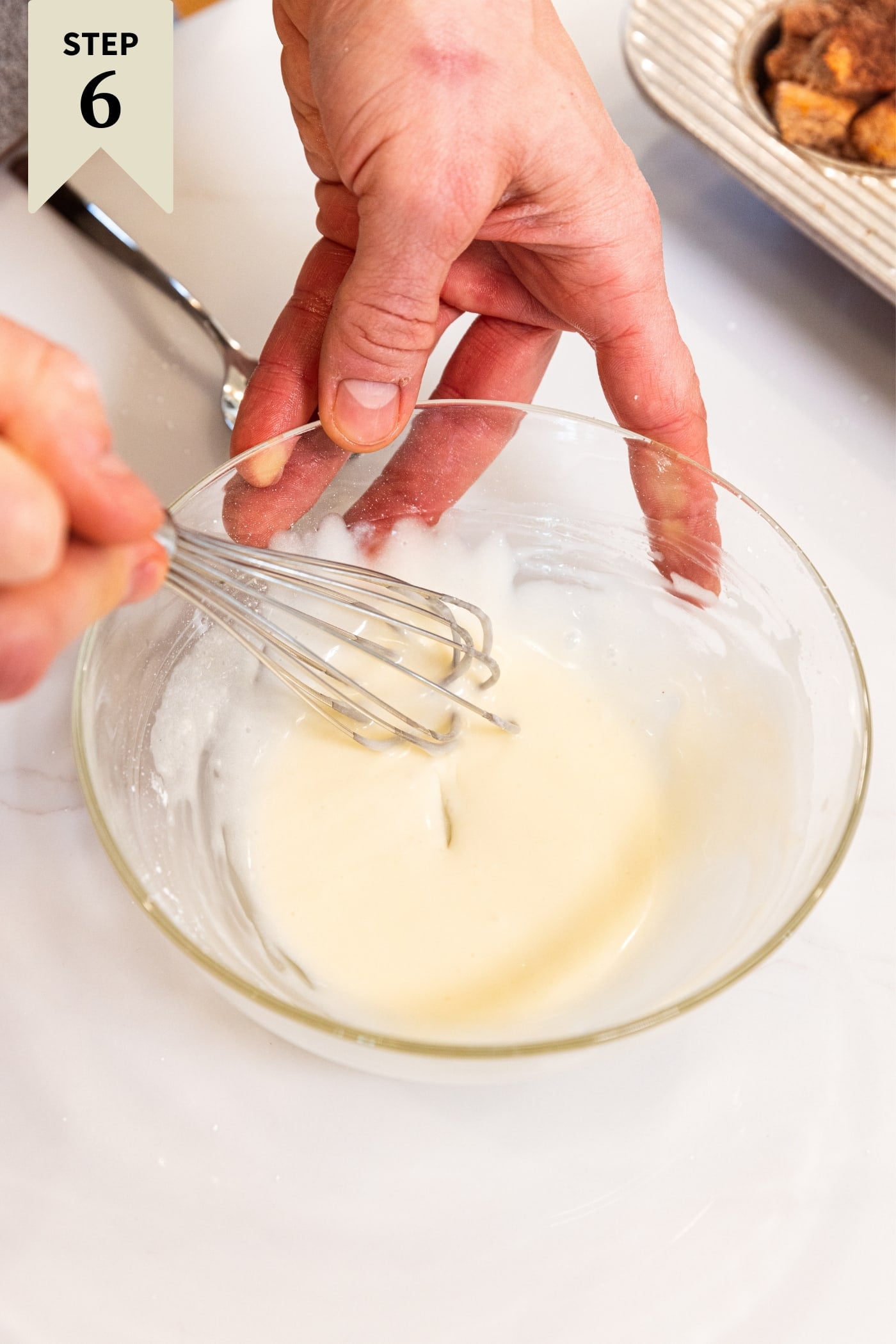 Hand mixing together white-colored thin sugar glaze in small glass mixing bowl on white countertop.