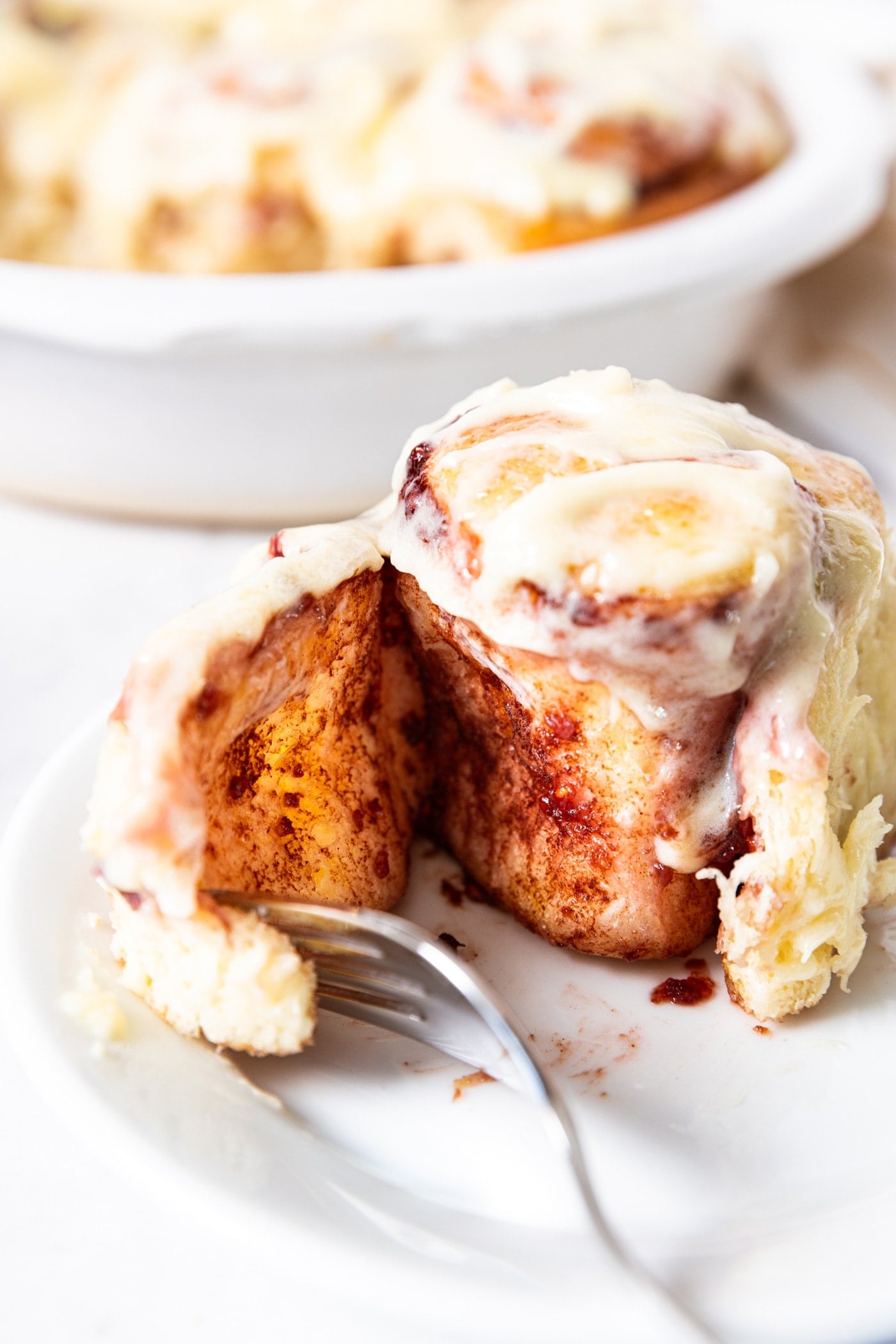 White plate with one raspberry cinnamon roll on it with fork pulling apart the roll showing the interior texture filled with raspberry filling and cinnamon.