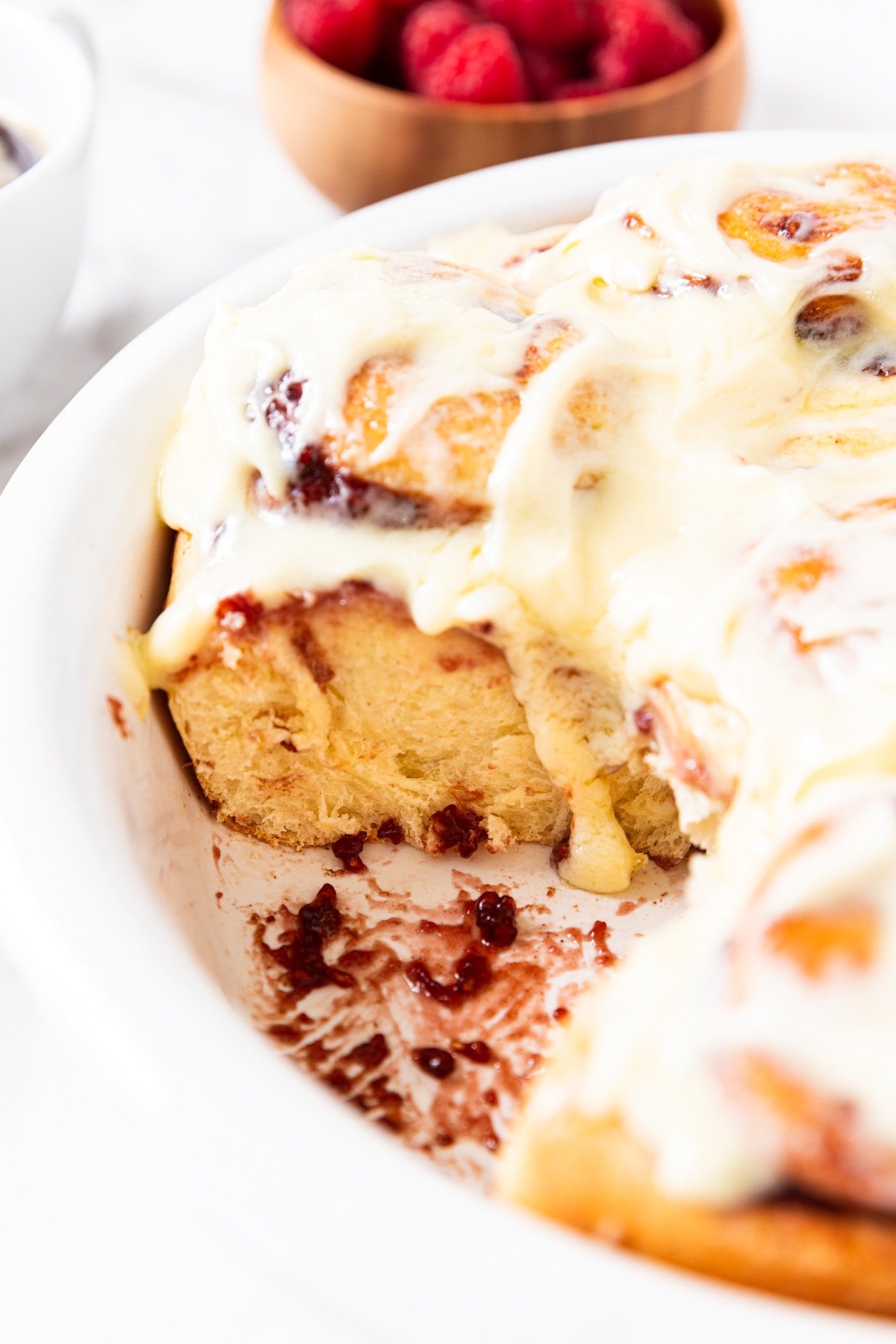Round baking pan filled with baked and frosted raspberry cinnamon rolls with one roll removed showing the texture of the cinnamon rolls with frosting dripping down.