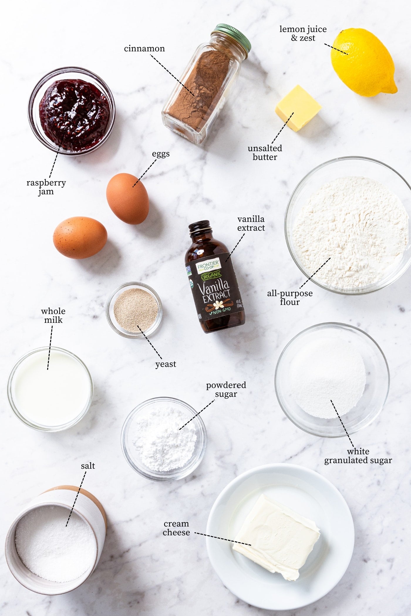 White marble surface filled with all ingredients needed to make raspberry cinnamon rolls including flour, sugar, butter, cinnamon, eggs, raspberry jam, cream cheese, and more.