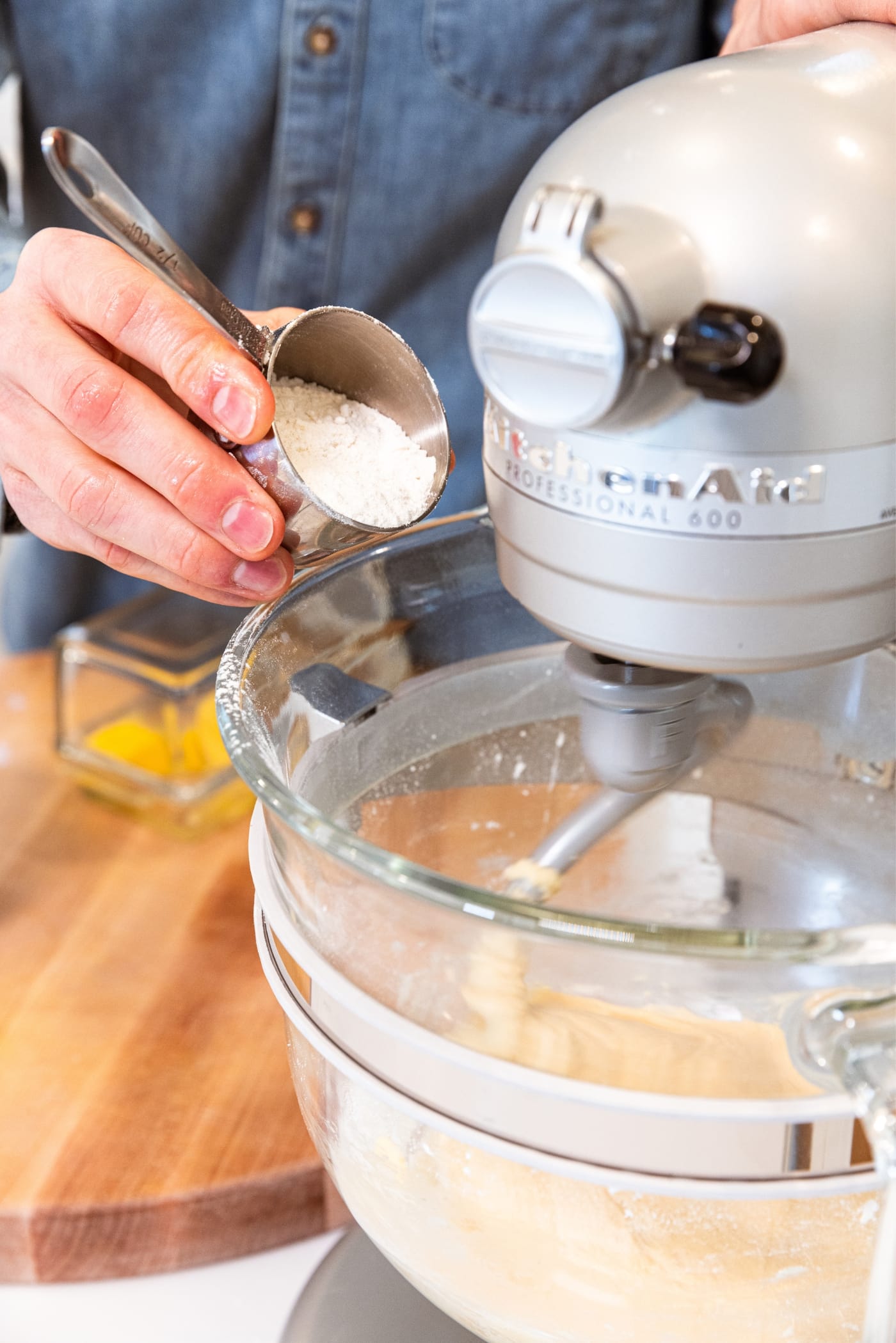 Hand holding small measuring cup pouring in flour into a large glass bowl attached to a stand mixer mixing together cinnamon roll dough.