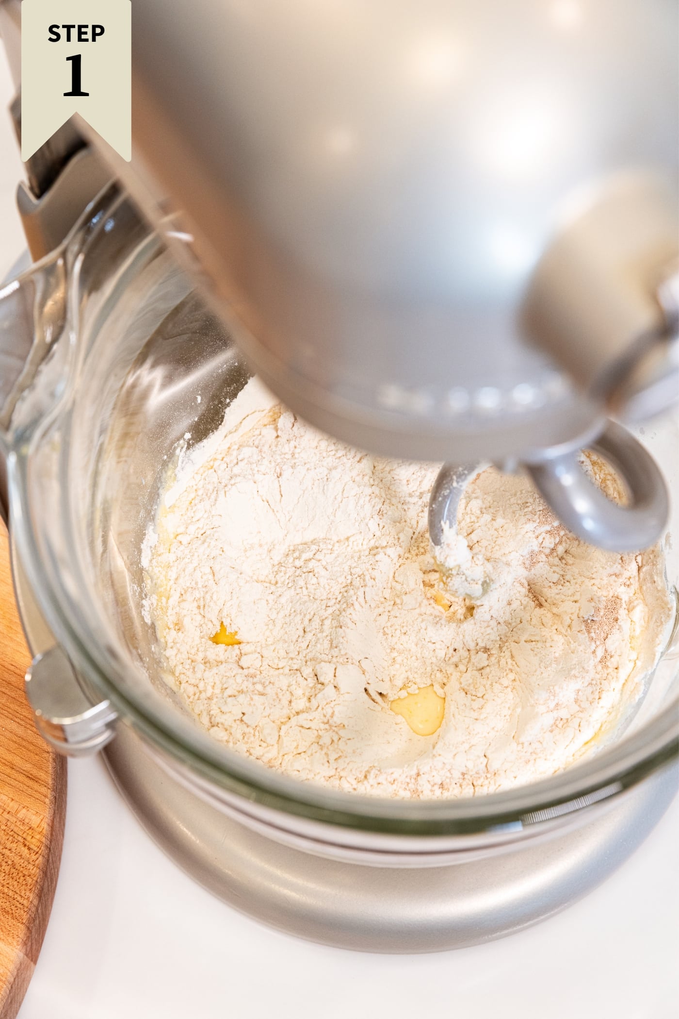 Stand mixer with glass bowl attached mixing together dry ingredients to make cinnamon rolls on white surface.